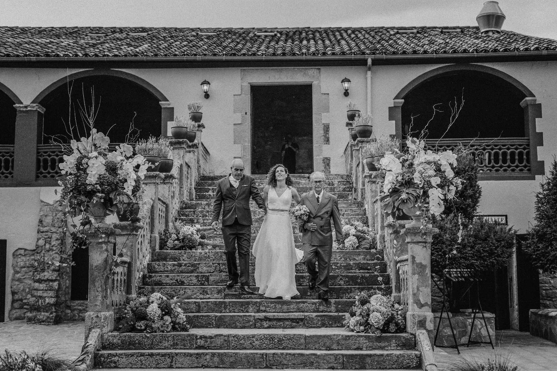 boda LGTBI en El Xaloc, Calders — la novia baja una gran escalera de piedra con dos familiares, en blanco y negro