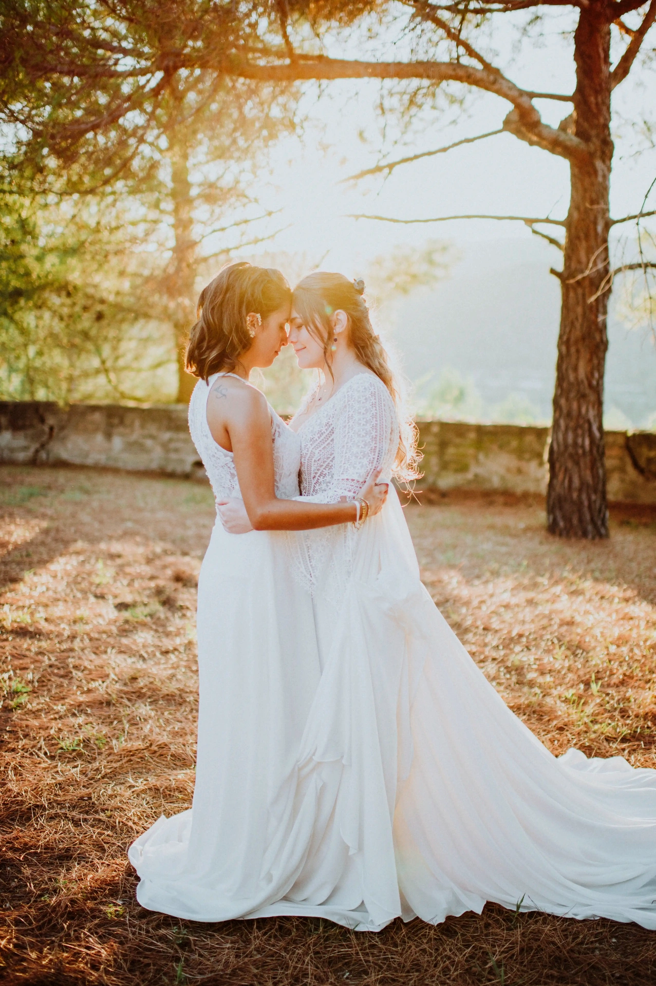 boda LGTBI en Ca n'Alzina — las novias frente con frente bajo los pinos con destellos de sol filtrándose entre las ramas