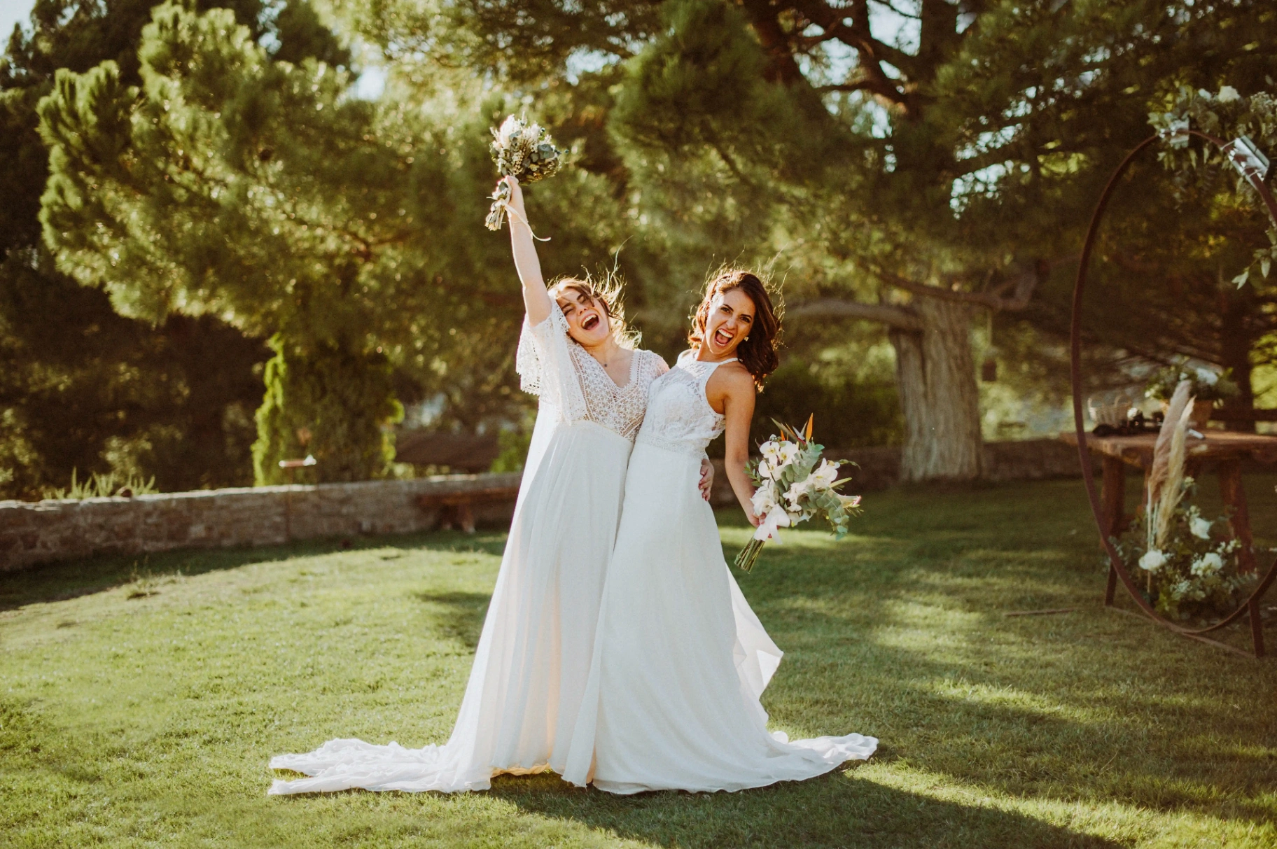 boda LGTBI en Ca n'Alzina (Rubió) — retrato de pareja de las novias celebrando con los ramos en alto entre pinos