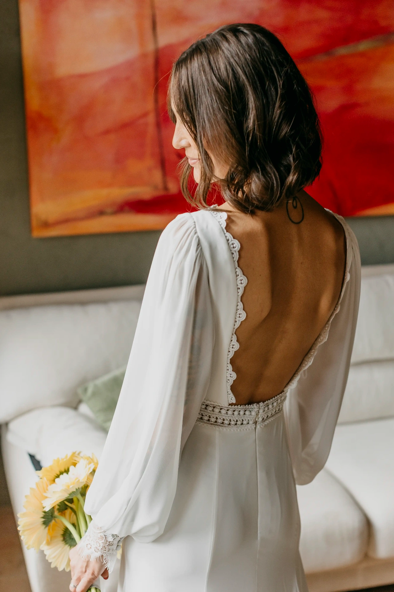 Retrato de espaldas de Carla con vestido de novia de espalda descubierta y ramo de gerberas amarillas frente a un cuadro rojo en Can Riera de la Pineda