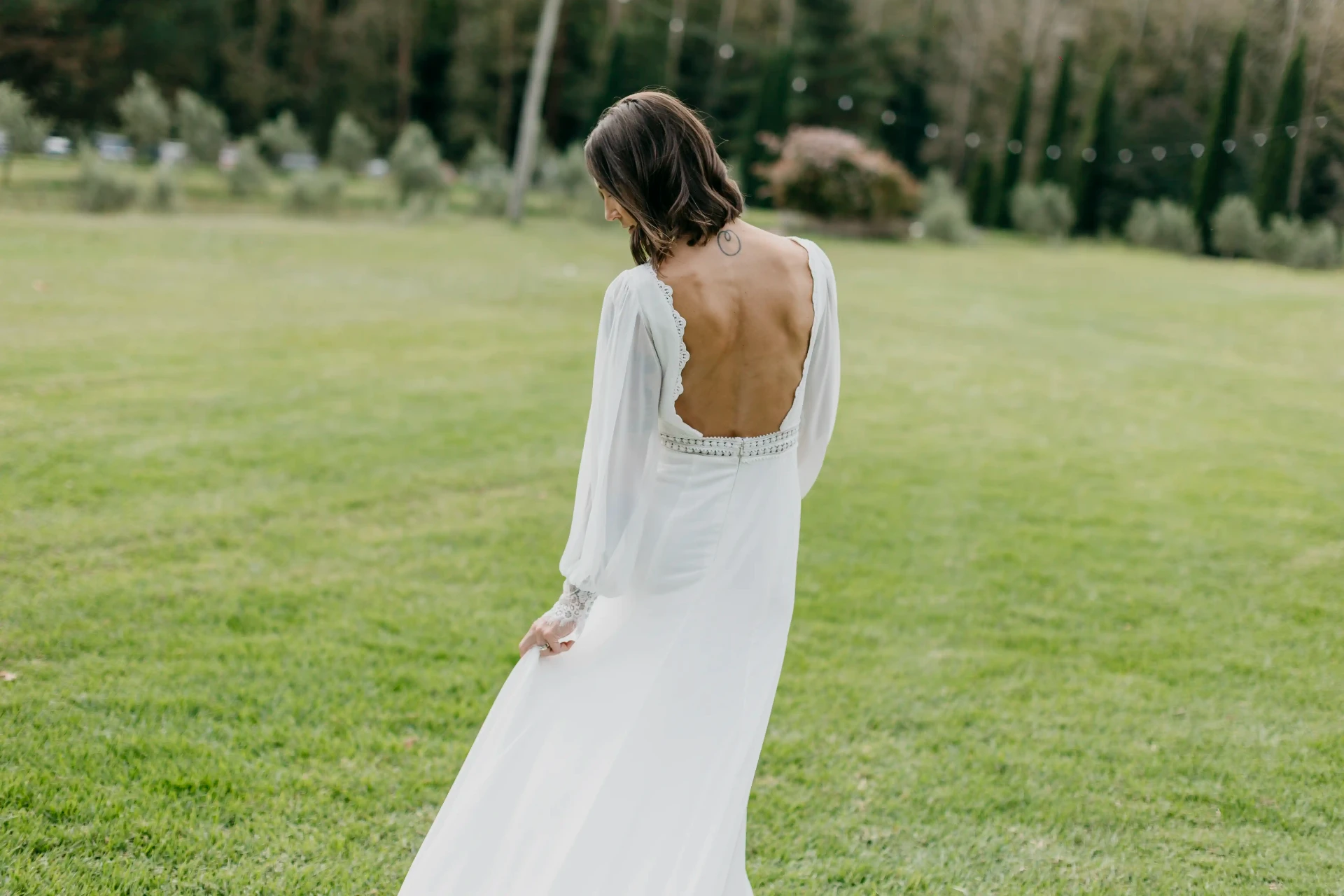Retrato de espaldas de Carla mostrando el detalle de la espalda descubierta de su vestido de novia en el jardín de Can Riera de la Pineda