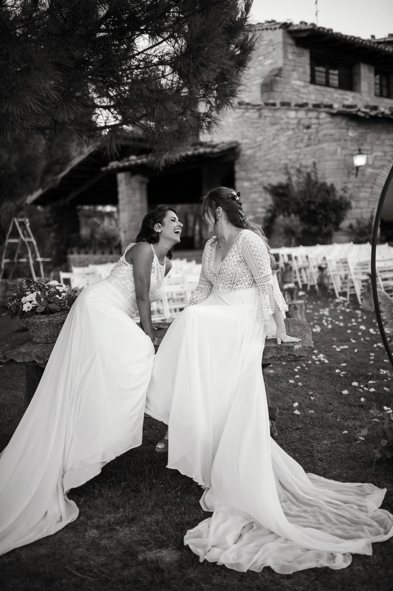 boda LGTBI en Ca n'Alzina — las novias riendo sentadas junto al altar en blanco y negro con la masía al fondo