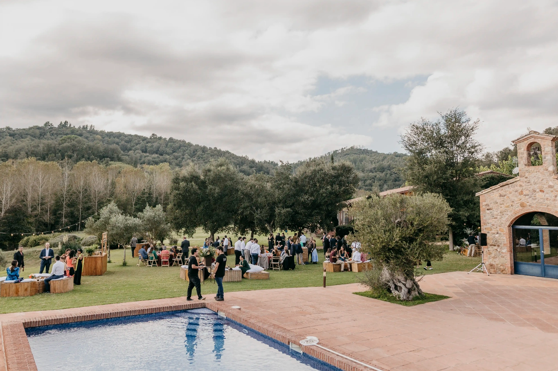Vista general del aperitivo al aire libre con invitados junto a la piscina y la ermita de piedra de Can Riera de la Pineda en el Montseny