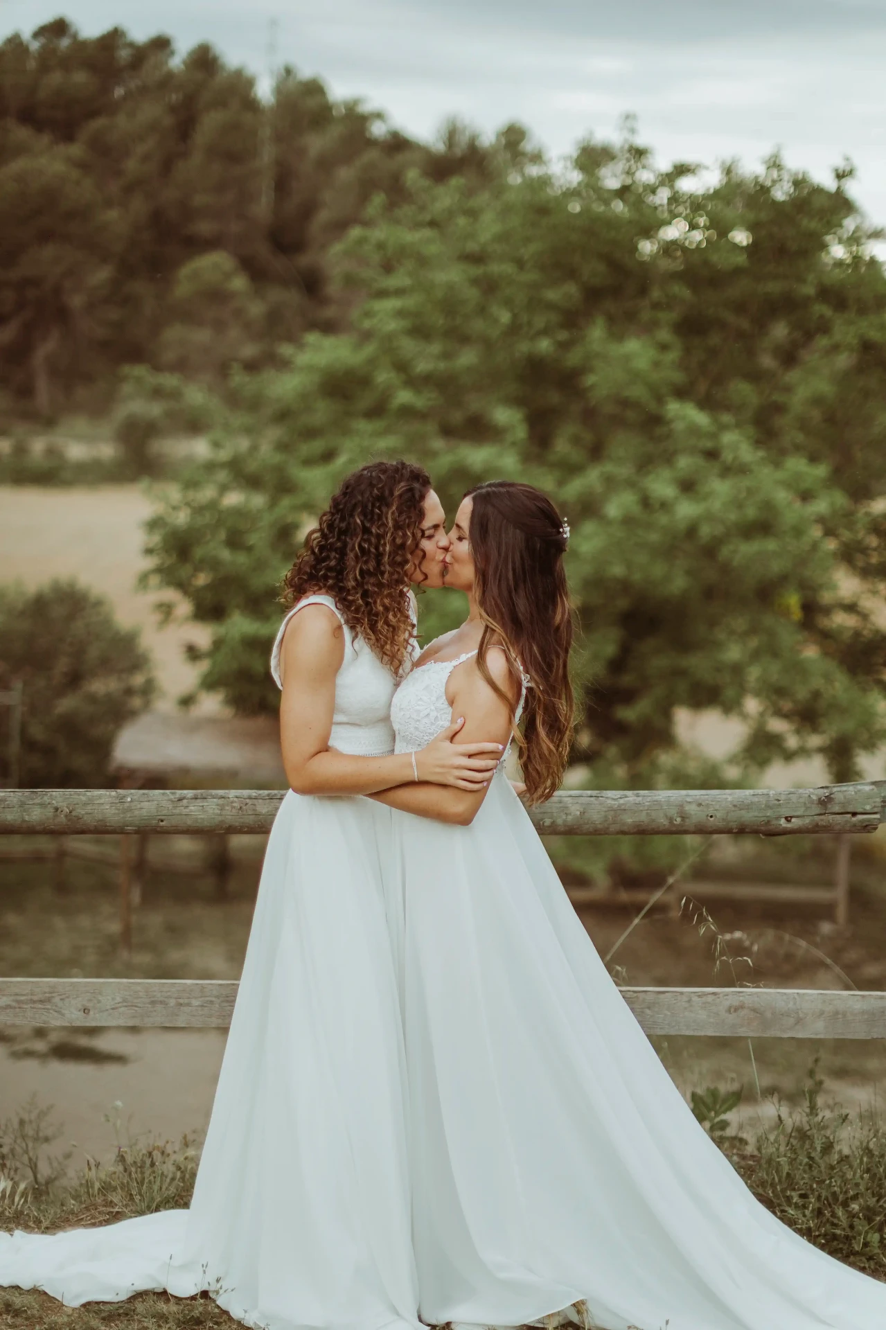 boda LGTBI en El Xaloc, Calders — beso de las novias frente a la valla de madera con los campos de fondo