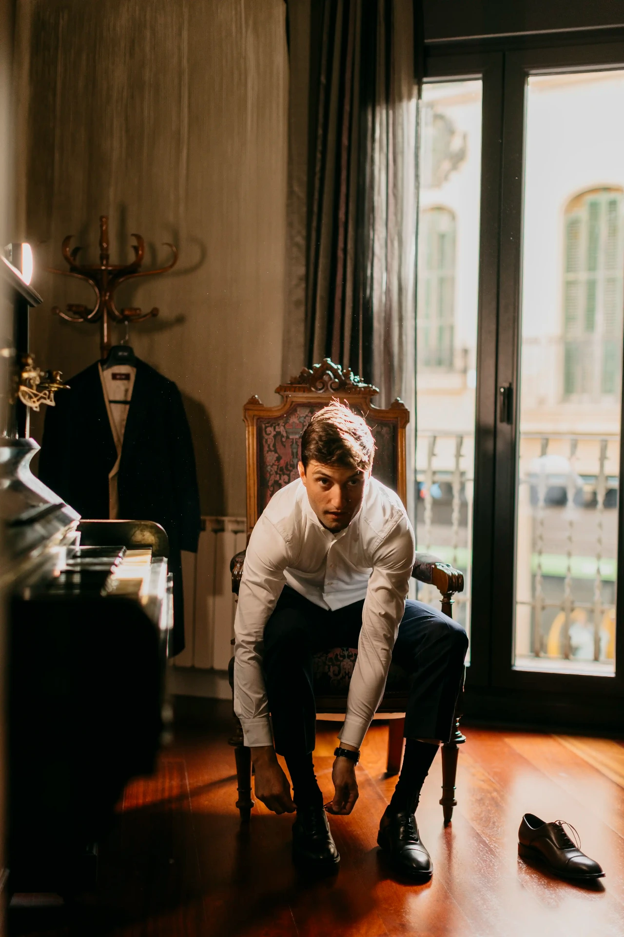 Novio atándose los zapatos junto a una silla clásica y ventana con luz natural, preparativos boda Lleida