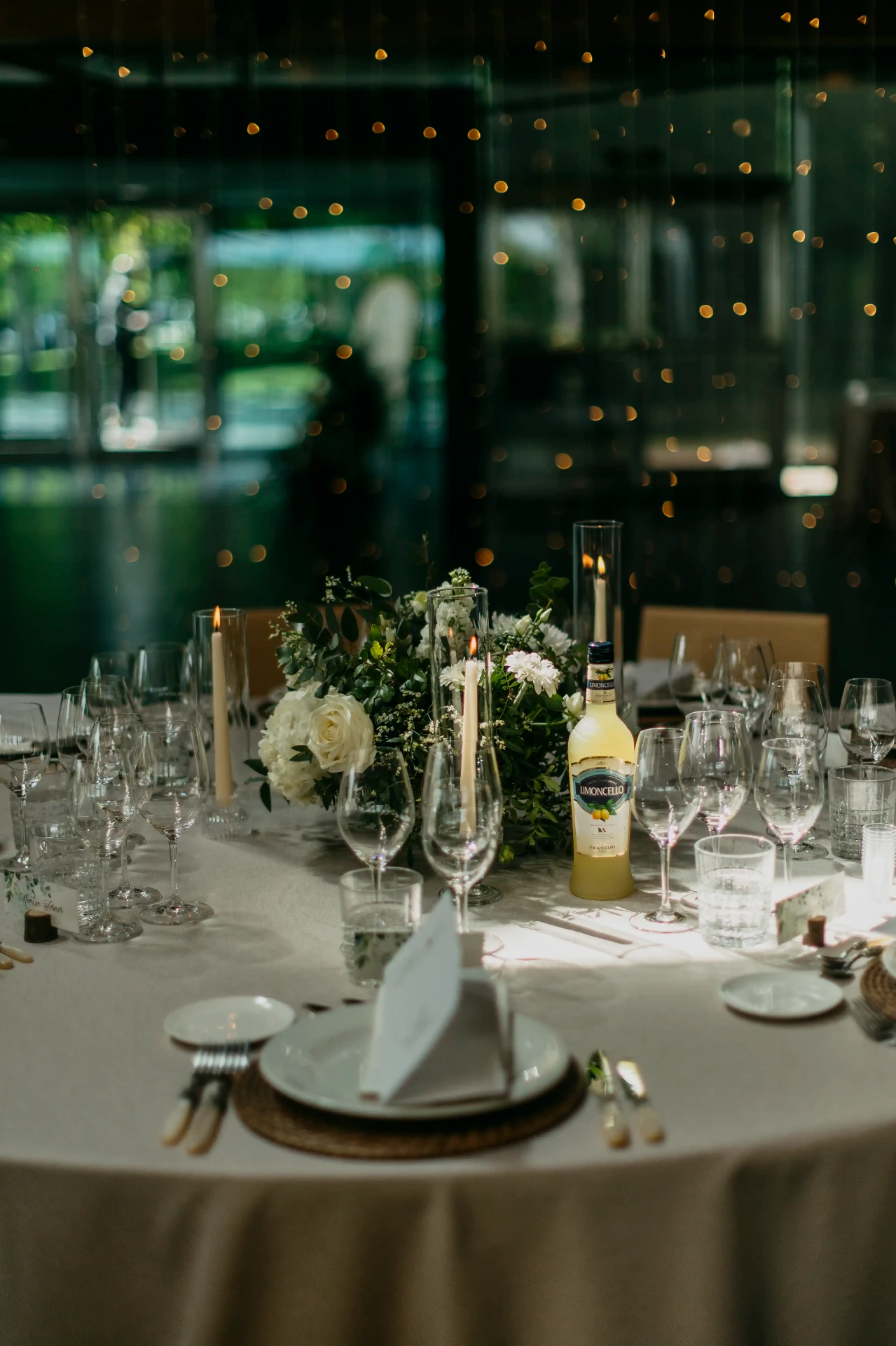 Mesa redonda del banquete con centro floral blanco, velas y botella de limoncello frente a cortina de luces, boda La Boscana Lleida