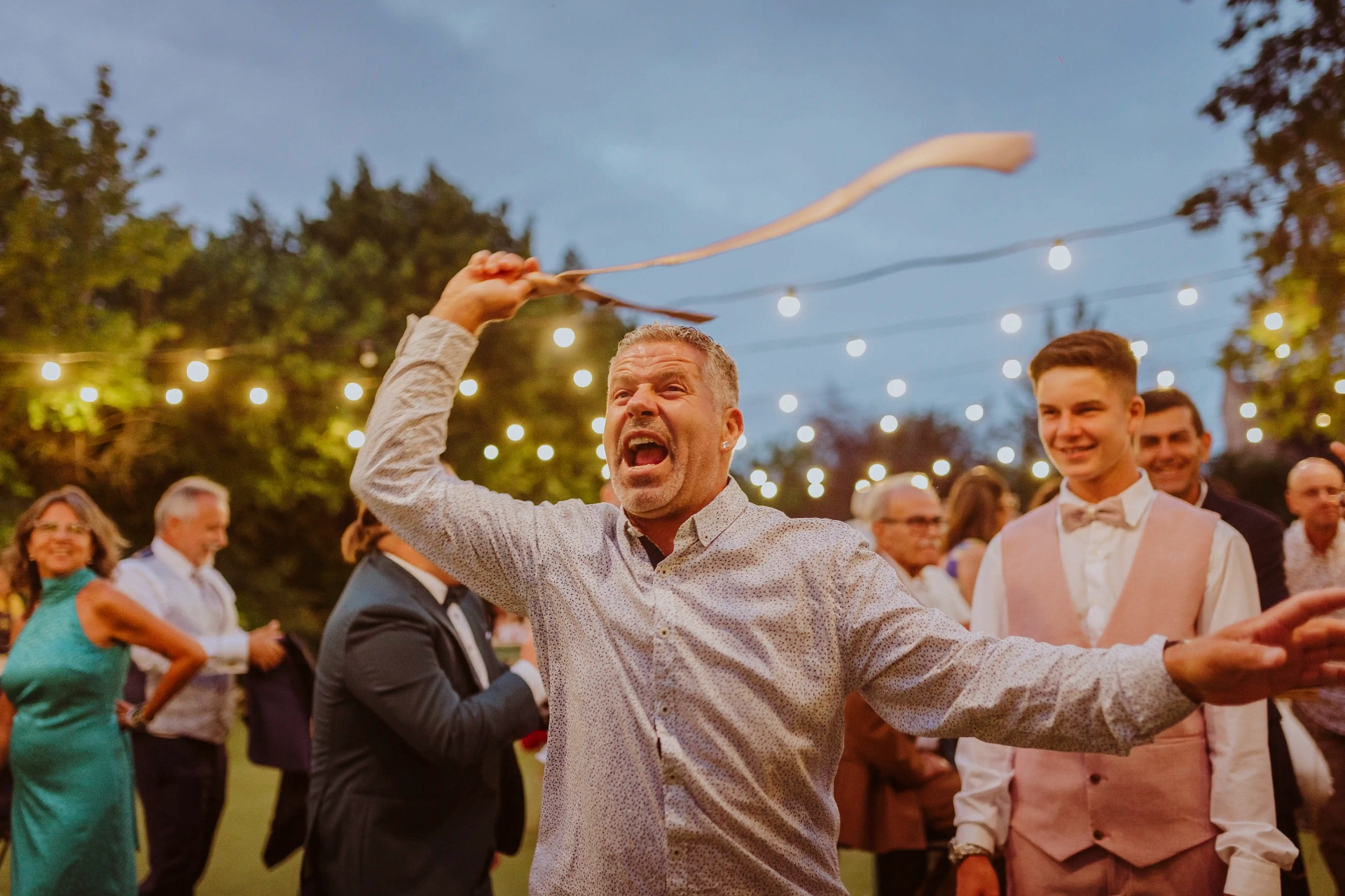 boda LGTBI en El Xaloc, Calders — invitado hace girar la corbata al aire riendo en la pista al atardecer