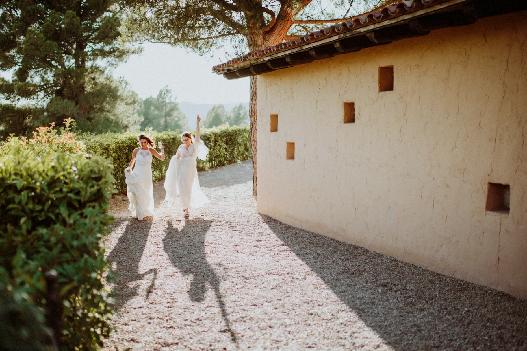 boda LGTBI en Ca n'Alzina — las novias caminando hacia el aperitivo por los jardines de la masía a última luz