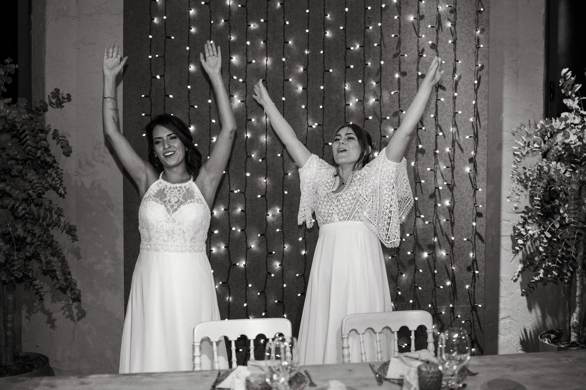 boda LGTBI en Ca n'Alzina — las novias con los brazos en alto celebrando en la fiesta con cortina de luces al fondo