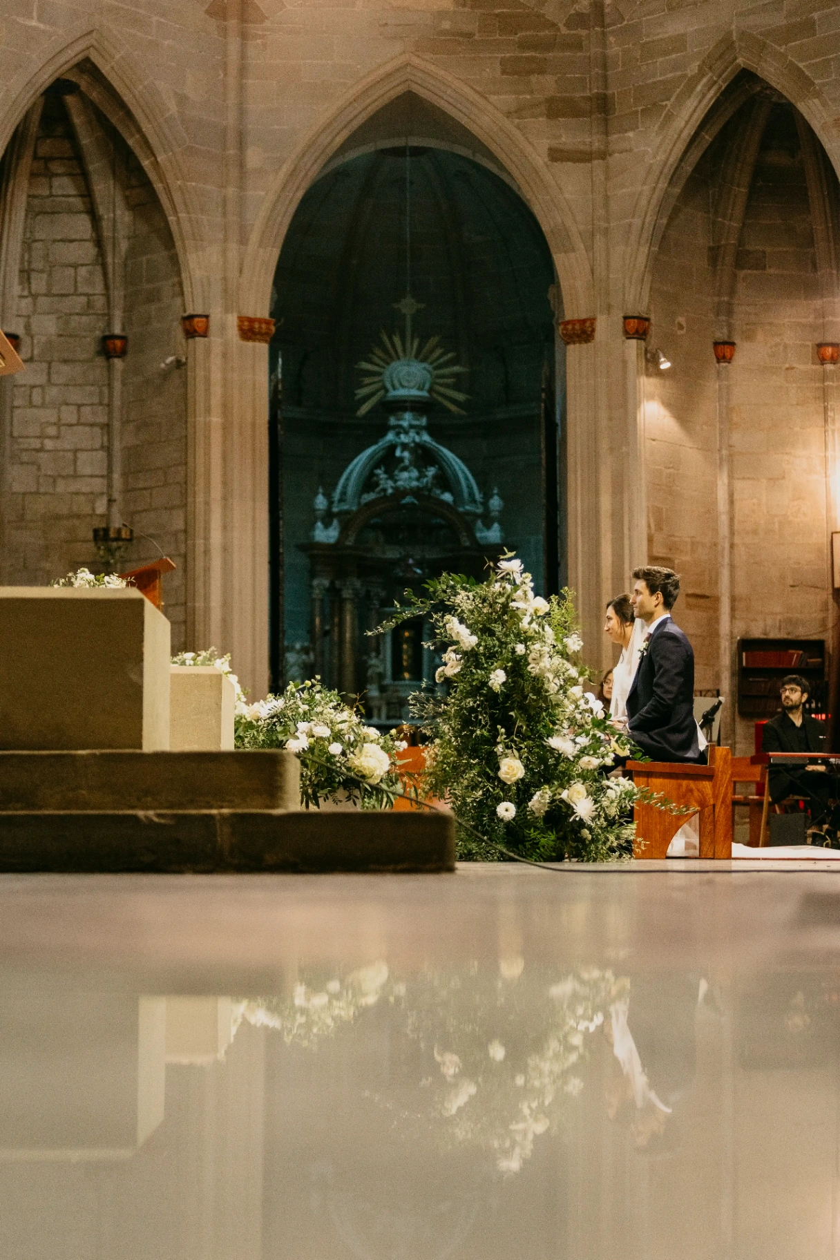 Novios sentados junto al altar con arreglo floral blanco y reflejo en el suelo, ceremonia boda en Cervera