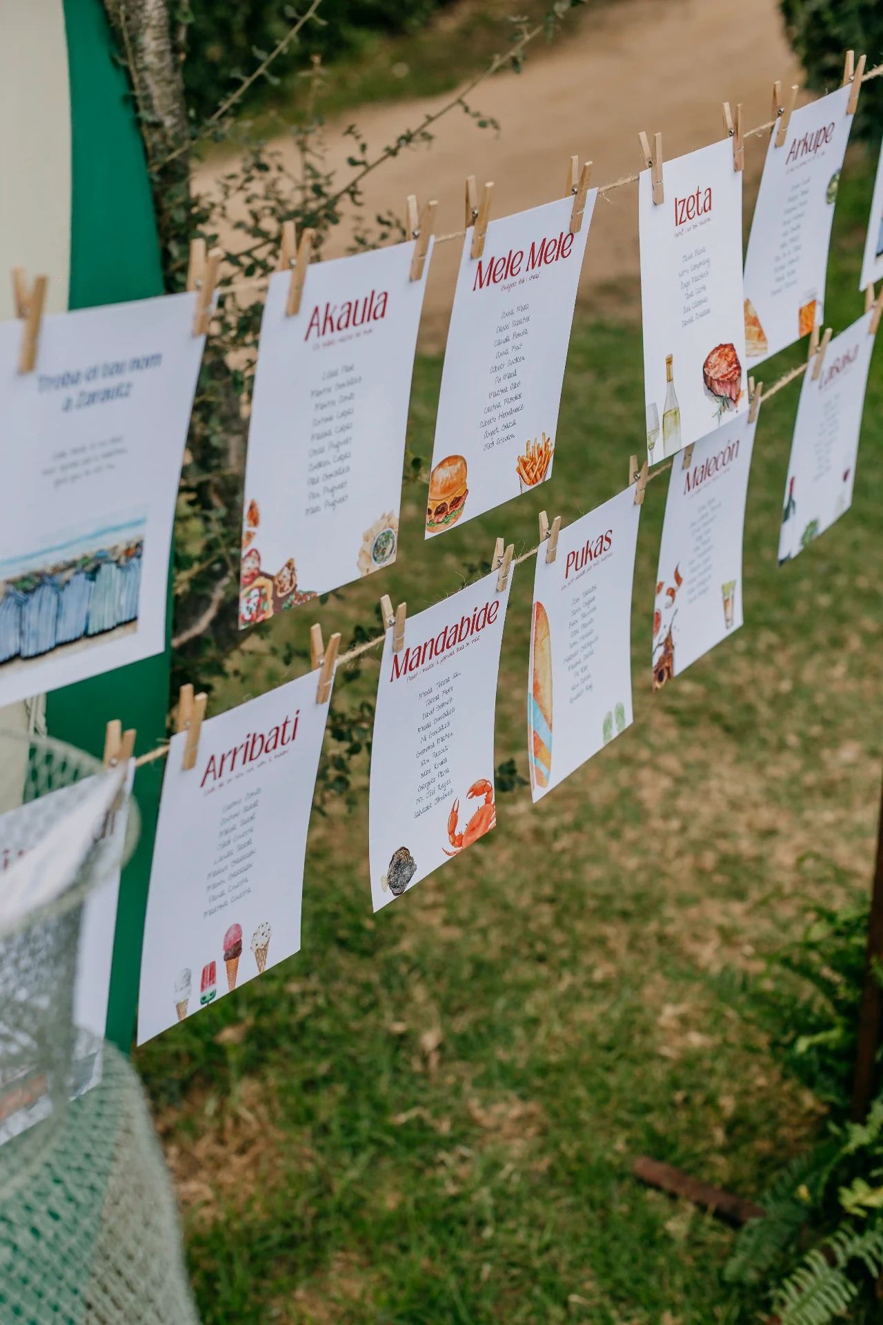 Seating plan artesanal con acuarelas de comida y nombres de mesas en euskera colgado con pinzas en el jardín de Can Riera de la Pineda