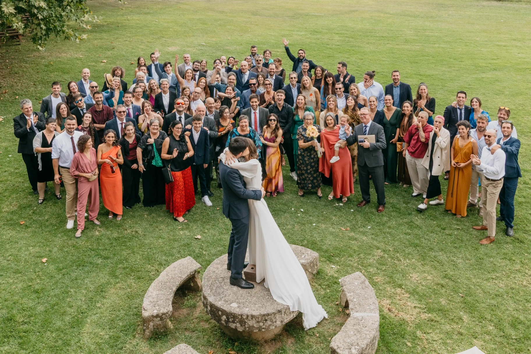 Foto de familia cenital con todos los invitados rodeando a Carla y Dani besándose sobre una piedra en el jardín de Can Riera de la Pineda
