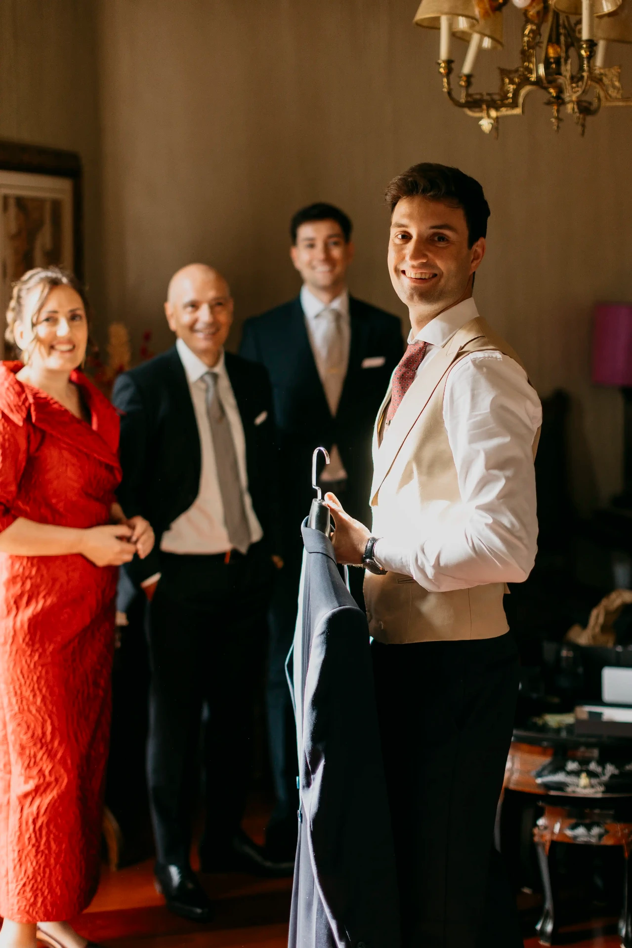 Novio sonriente con chaleco sujetando la americana rodeado de familia en los preparativos, boda en Lleida
