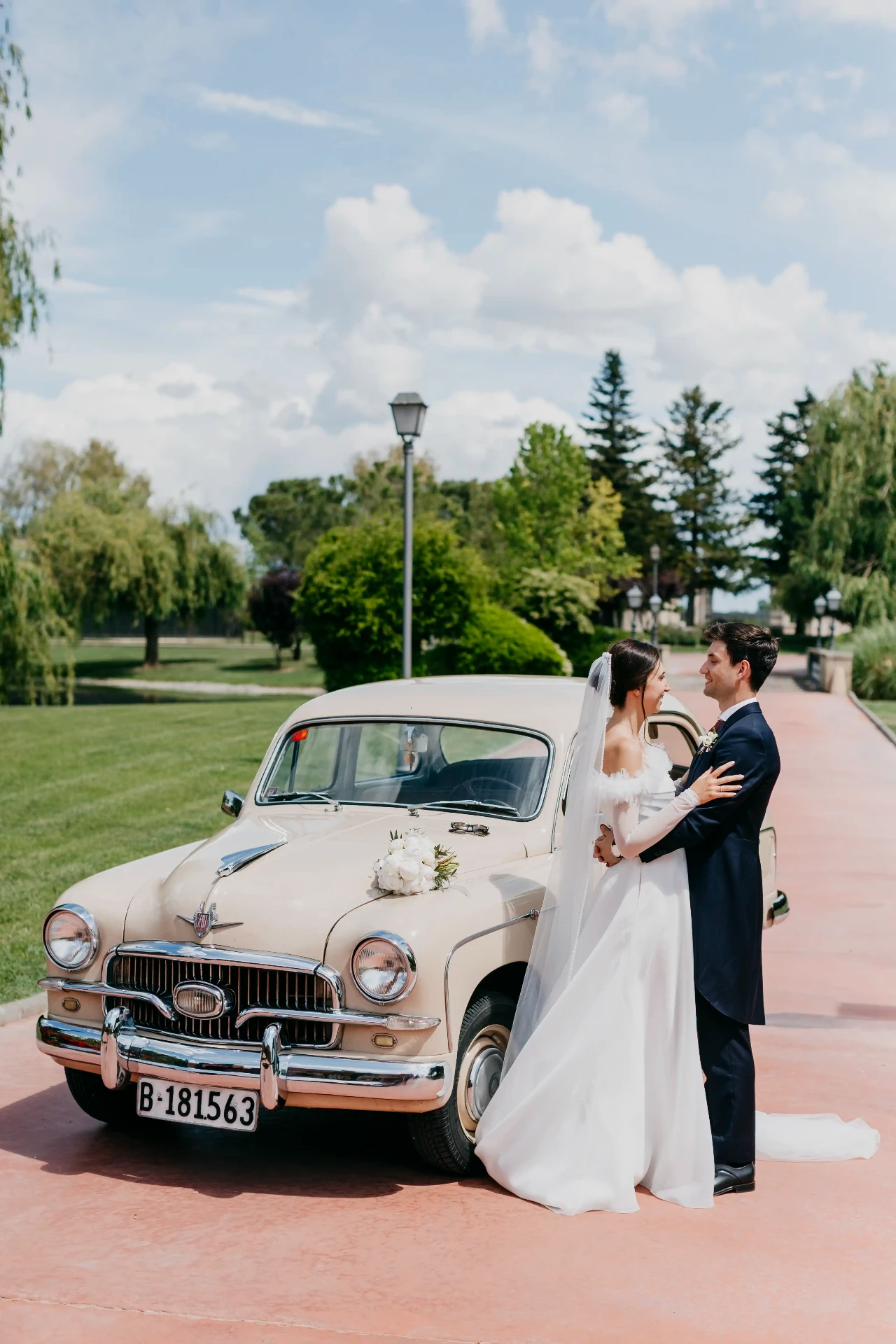 Joan y Núria abrazados junto al coche clásico decorado con flores en los jardines de La Boscana, boda Bellvís Lleida