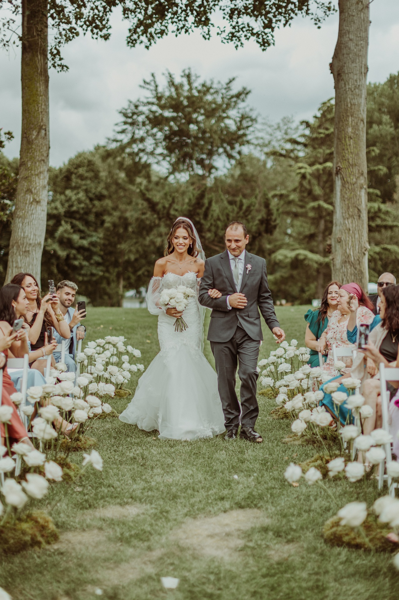 Alba entrando del brazo de su padre por el pasillo de rosas blancas en la ceremonia al aire libre entre los árboles