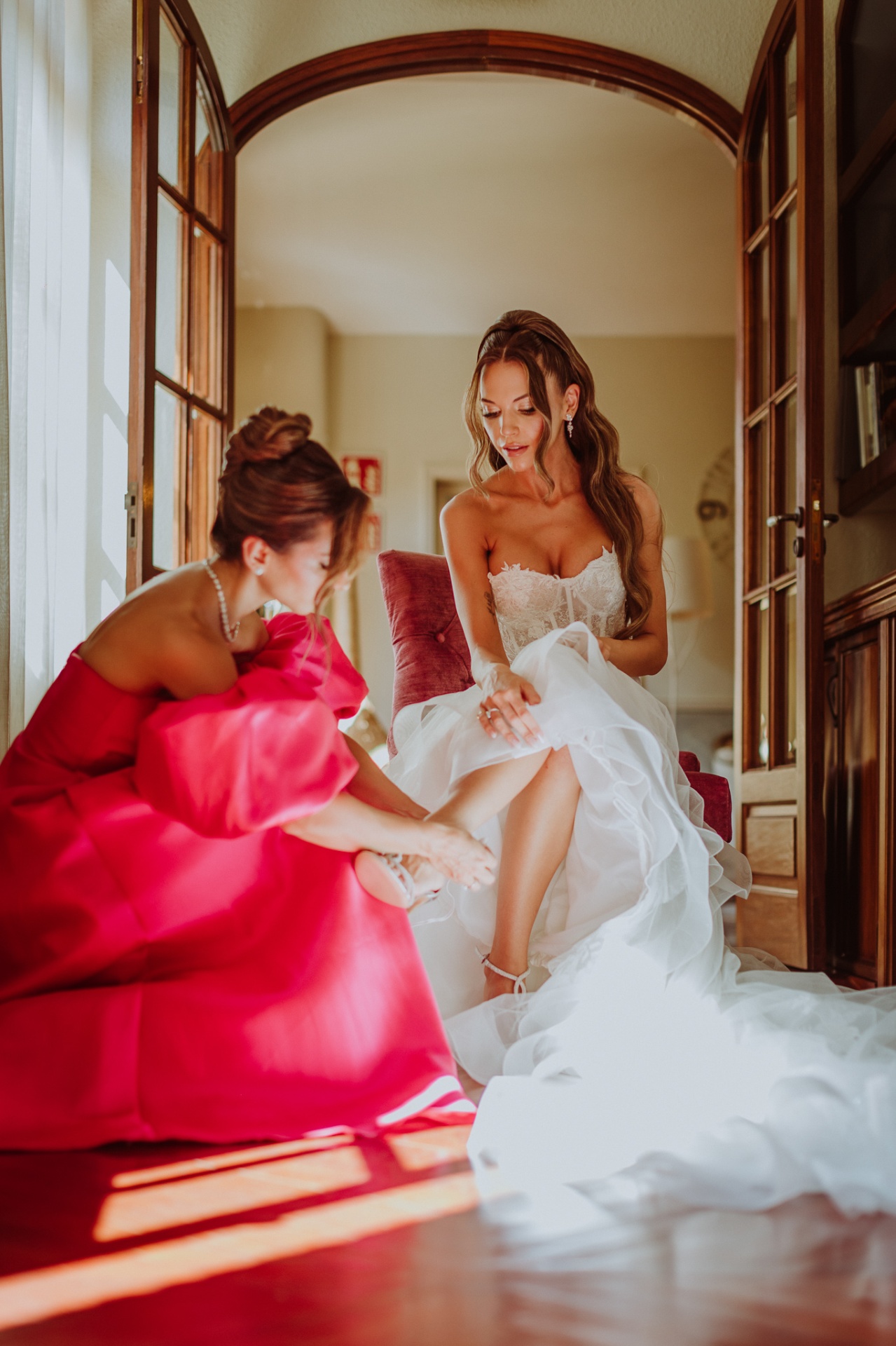 Alba sentada mientras su madre le ayuda a ponerse los zapatos de novia con luz natural entrando por las ventanas