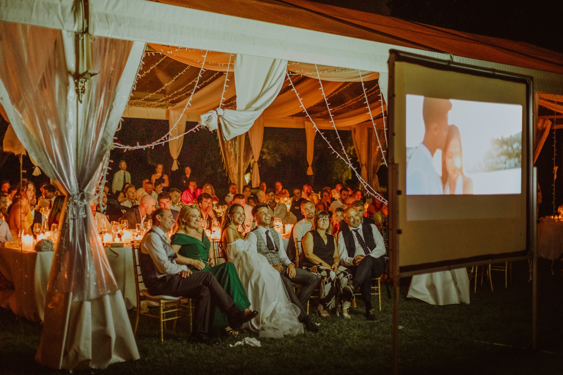 Alba y Roger viendo el same day edit en pantalla grande con los invitados bajo la carpa iluminada con velas