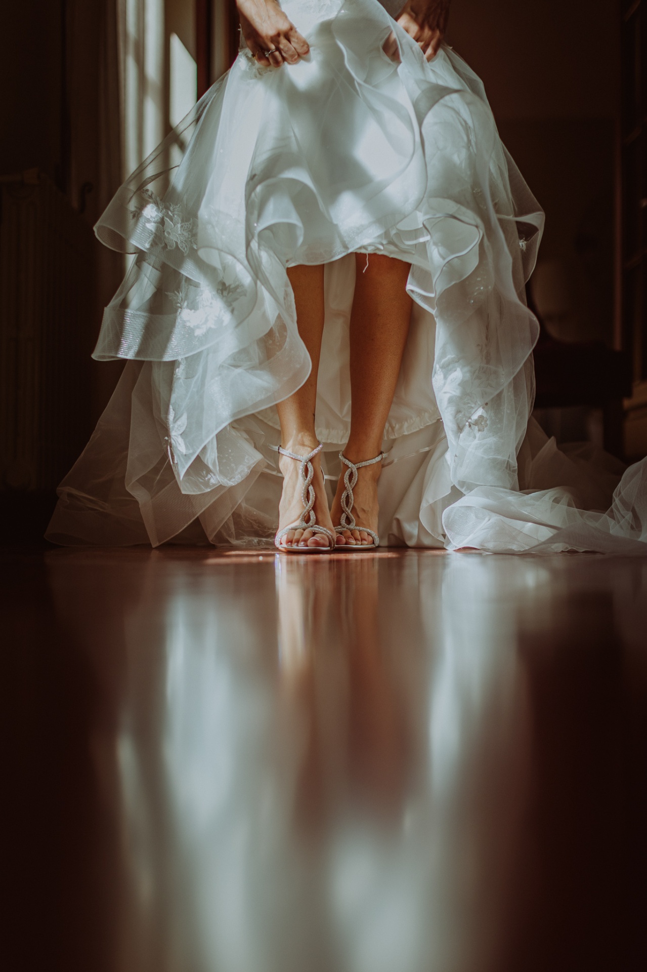 Detalle de los zapatos de novia con tiras trenzadas de pedrería y la falda de tul del vestido reflejados en el suelo