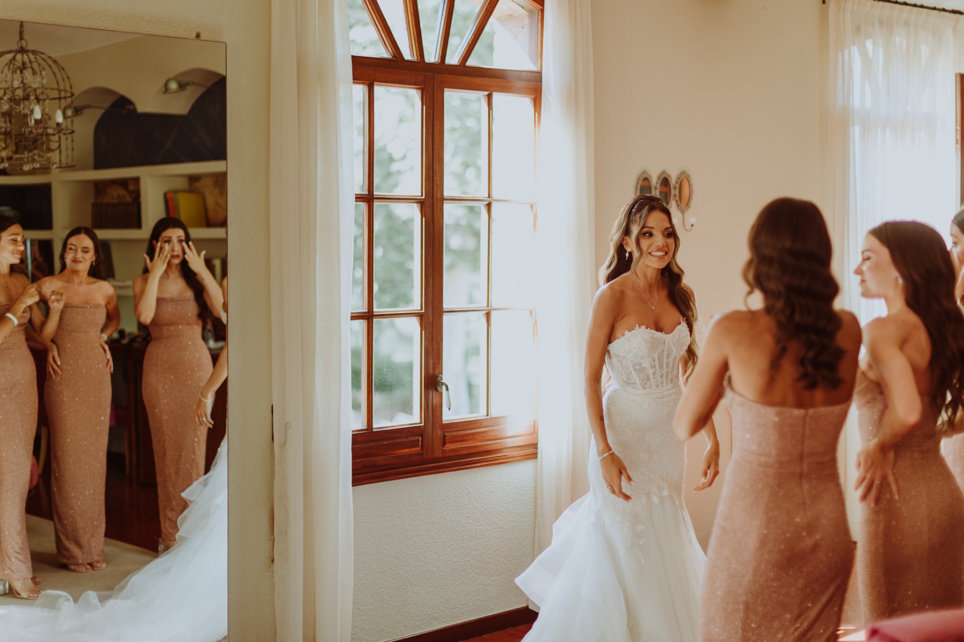 Alba sonriendo radiante con su vestido de novia mientras las damas de honor emocionadas se miran en el espejo