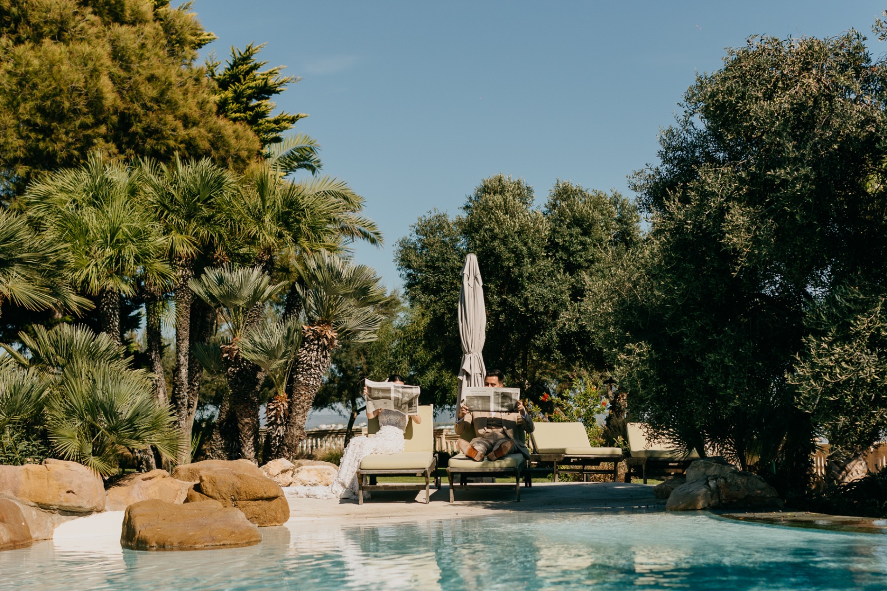 Groomsmen reading newspapers by the pool at Xalet del Nin surrounded by palm trees