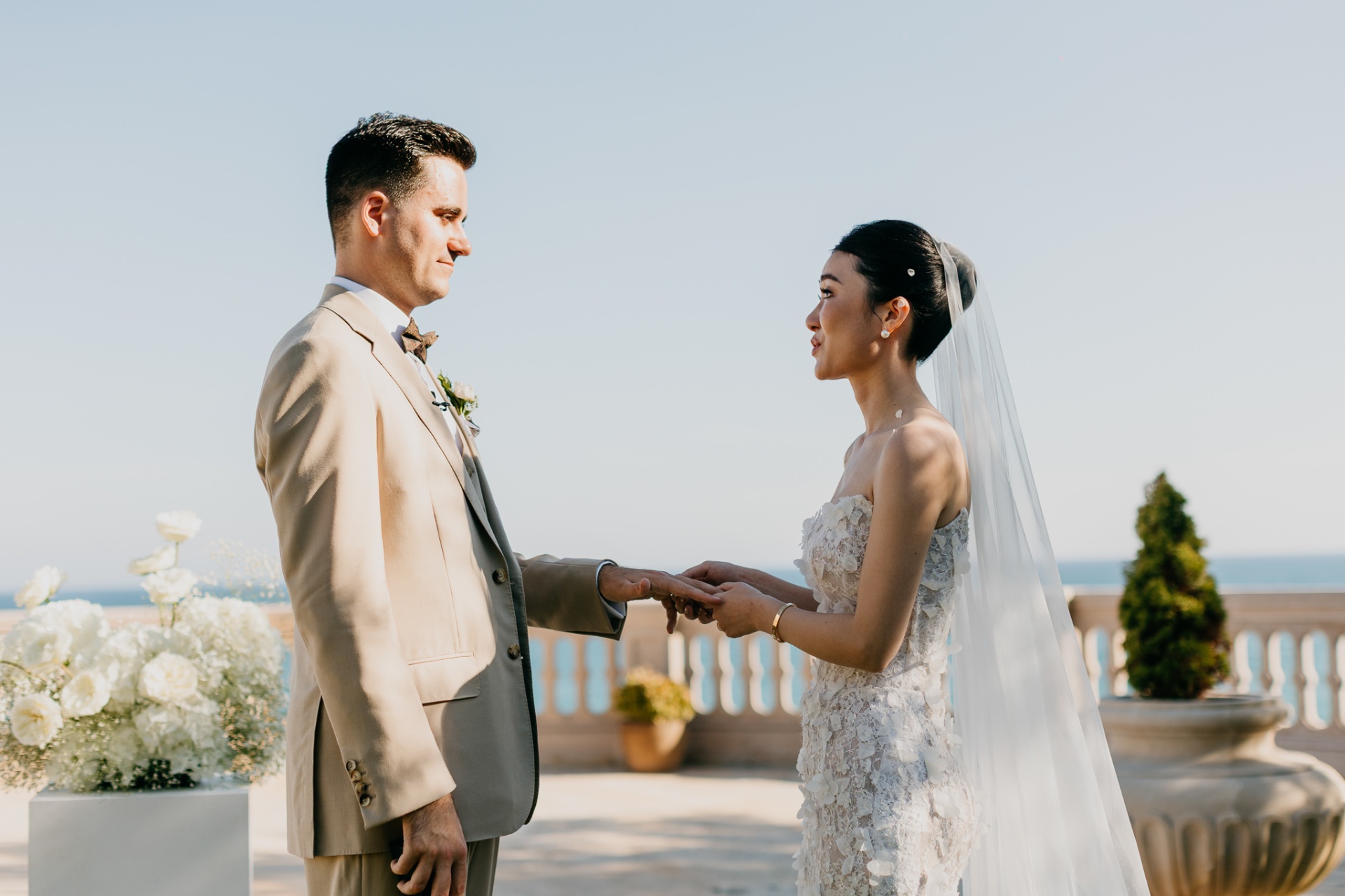 Cameron and Kara exchanging rings with the Mediterranean Sea and white floral arrangements in the background