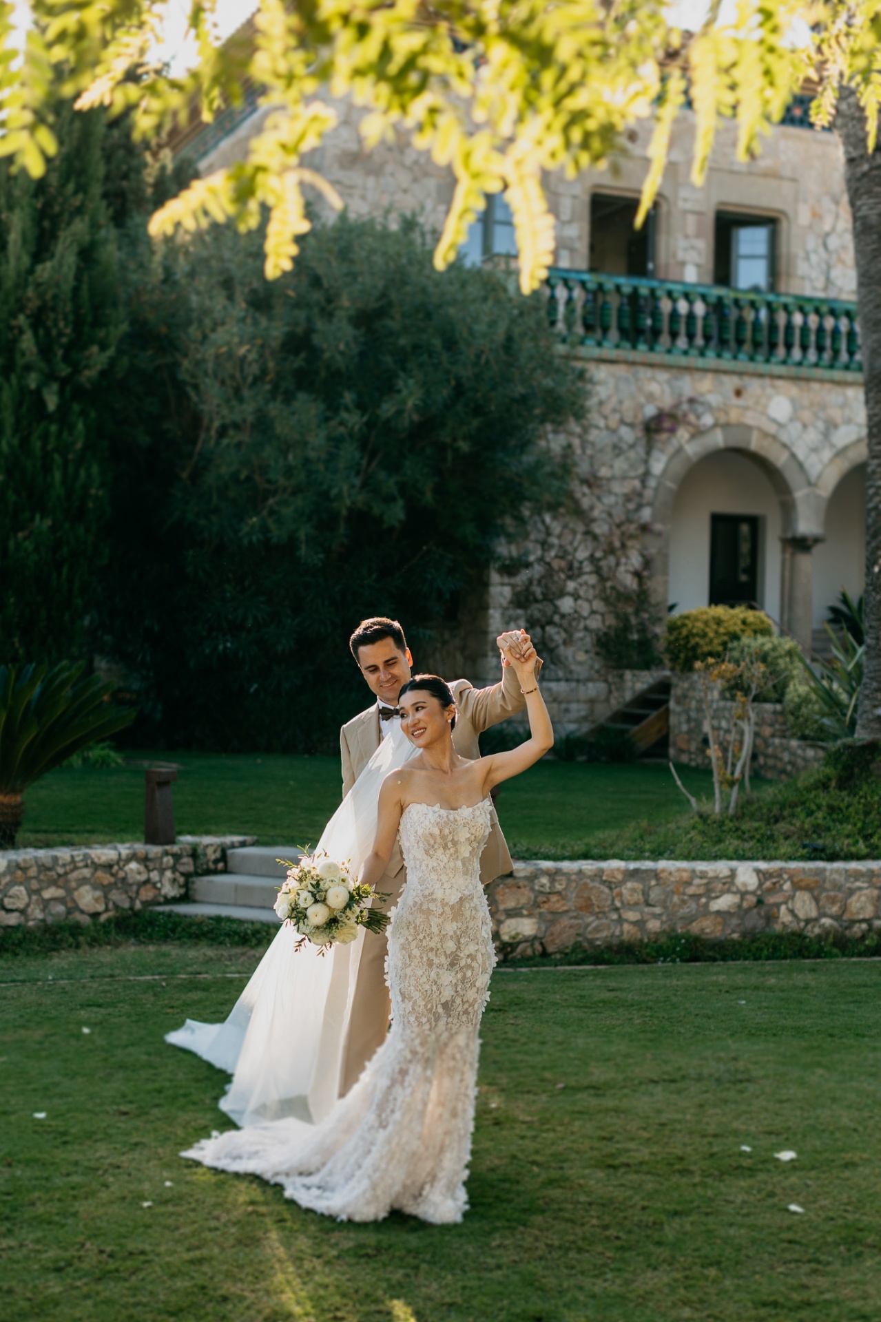 Cameron and Kara celebrating with raised hands and bouquet on the garden lawn of Xalet del Nin