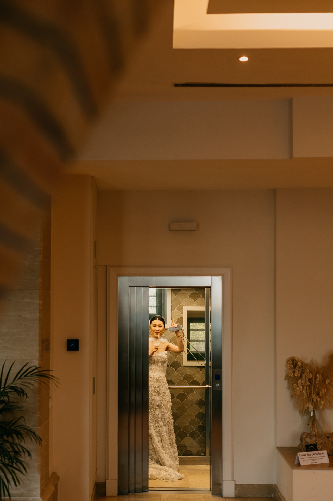Kara waving from an elevator in her wedding dress with a cocktail in hand at Xalet del Nin