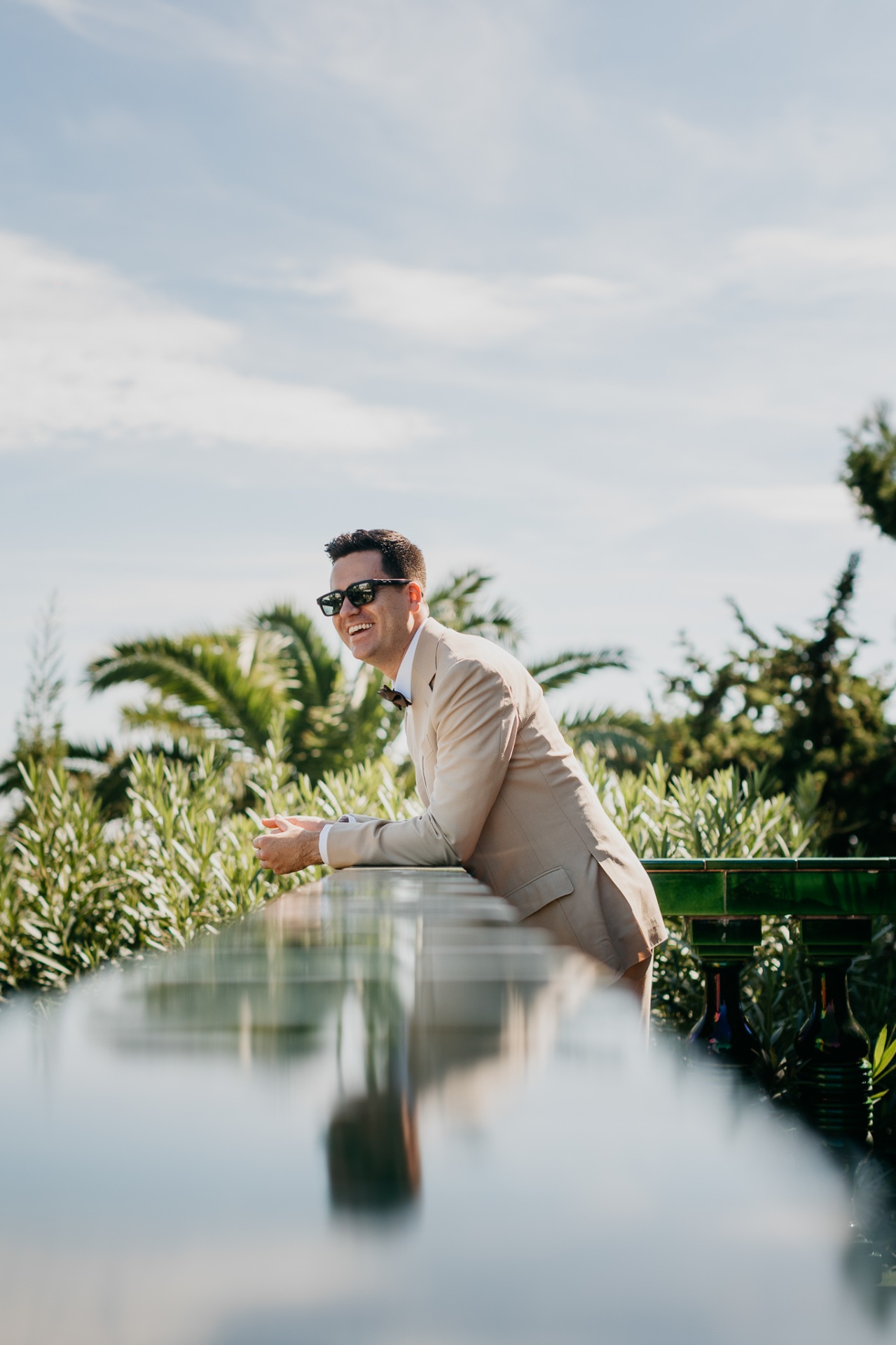 Cameron smiling in sunglasses leaning on the glazed ceramic balustrade with palm trees behind