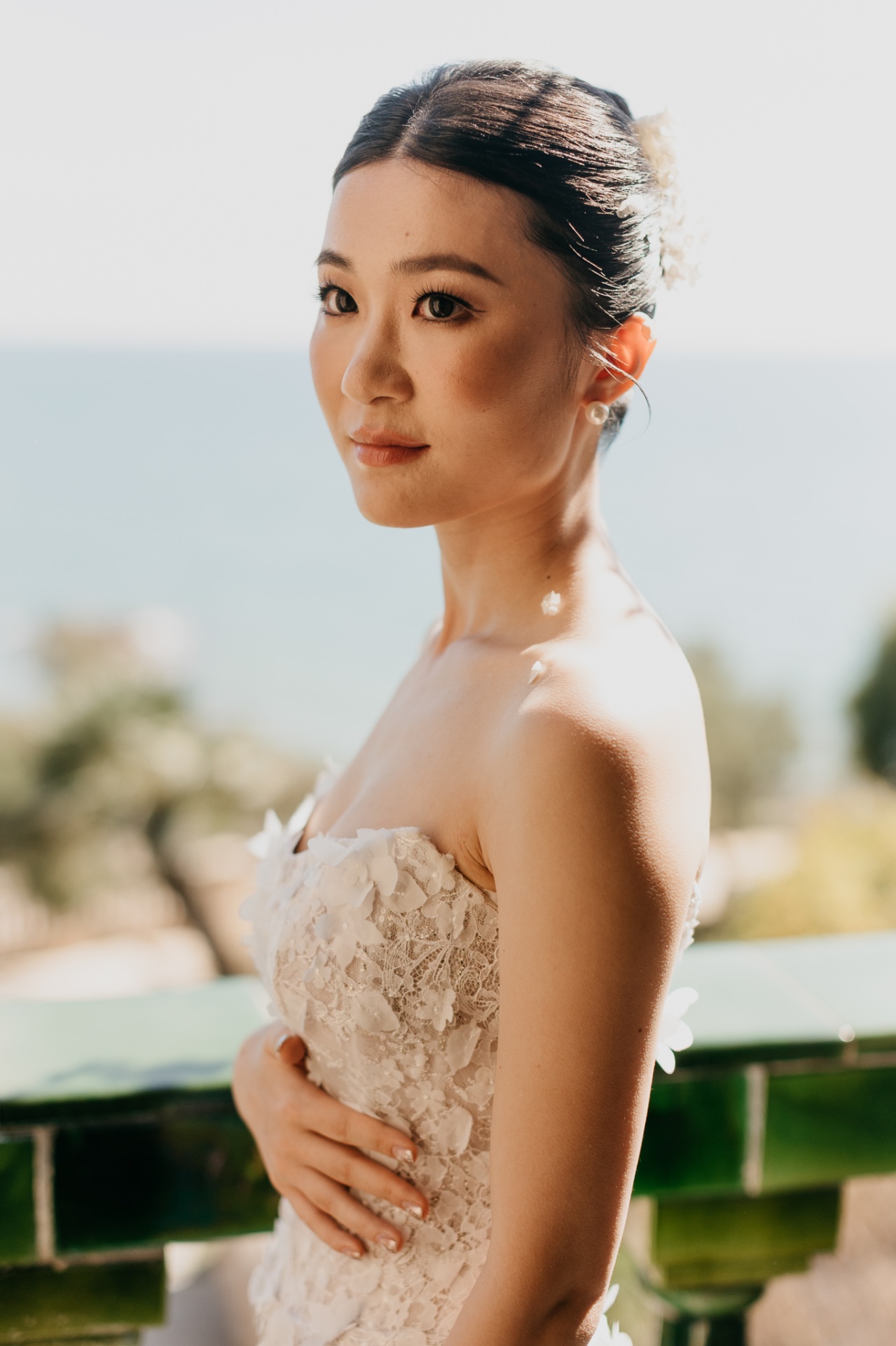 Close-up portrait of Kara with pearl earrings and floral hair accessory against the sea at Xalet del Nin
