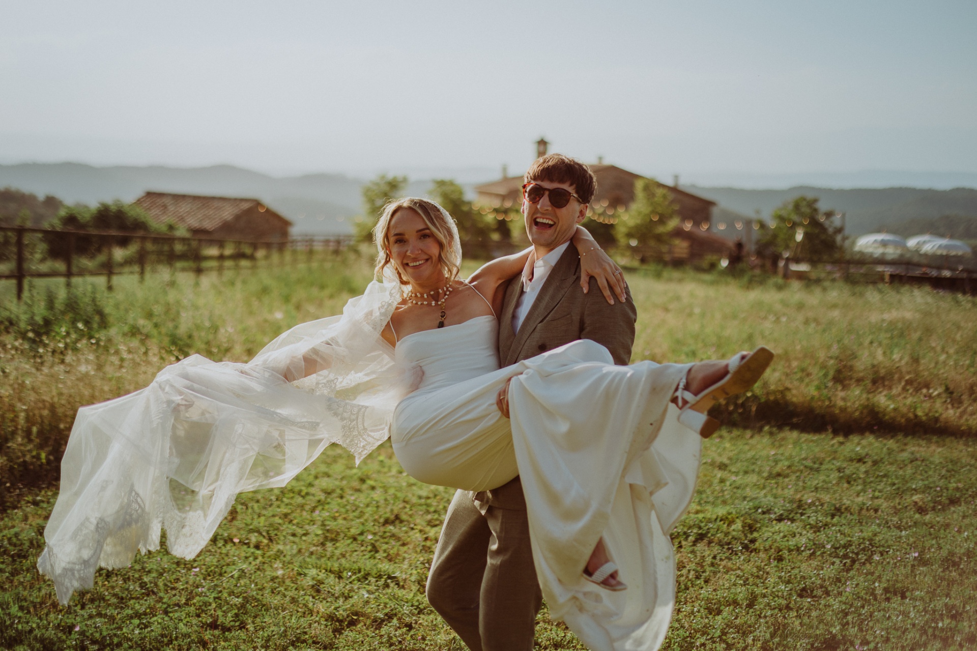 Conor carrying Ellie laughing in his arms at sunset with masía behind