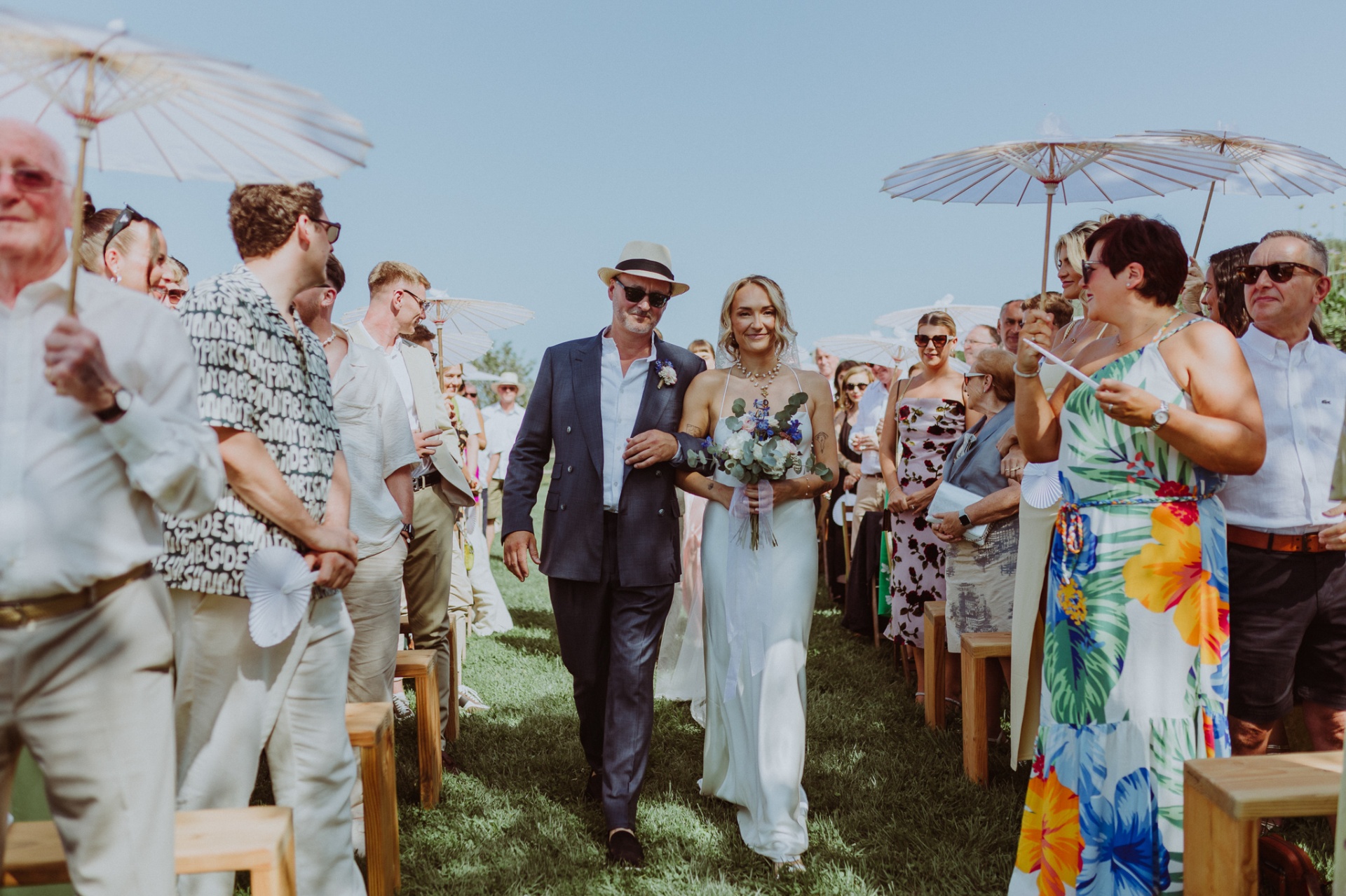 Ellie walking down the aisle with her father among guests with parasols
