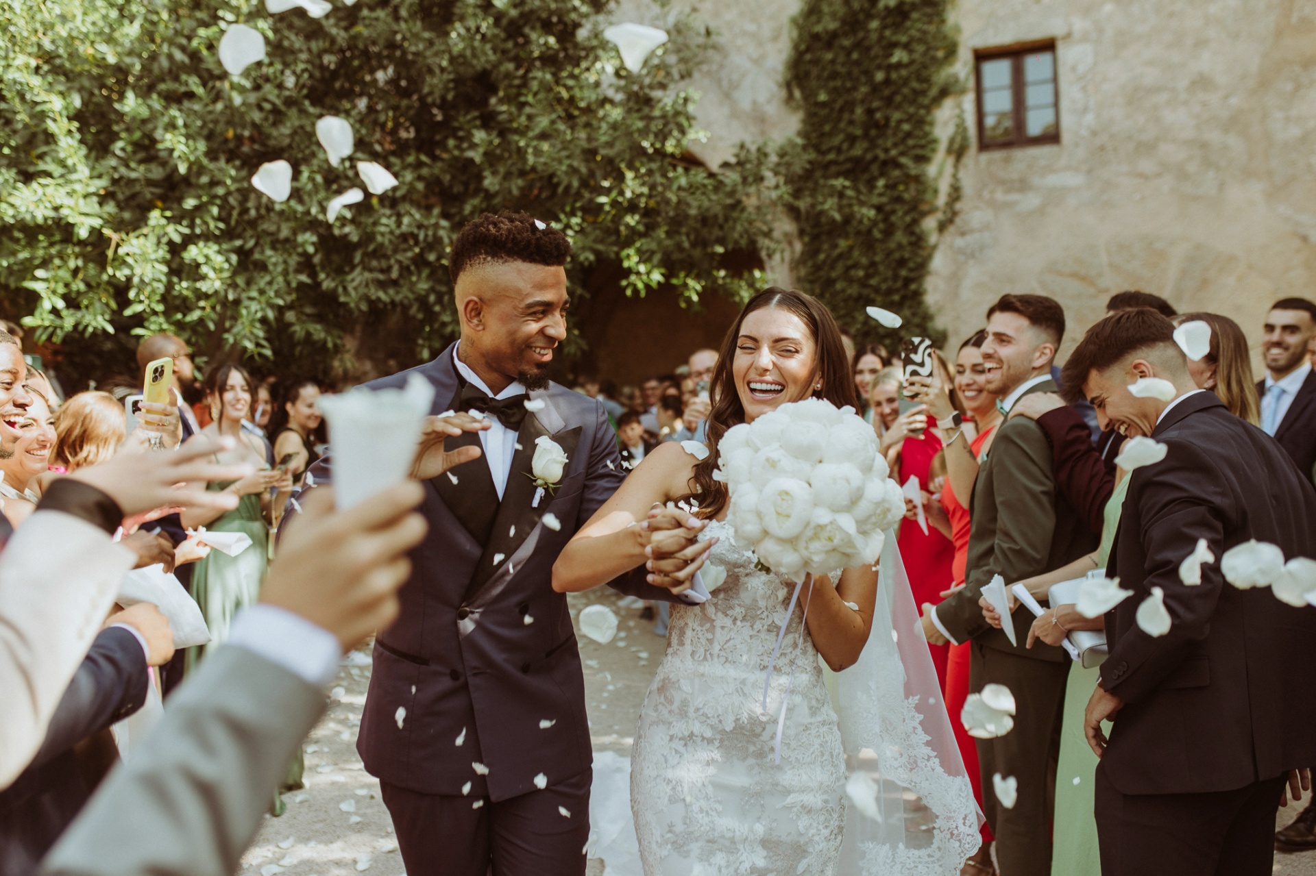 Laura and Jose laughing under white petal confetti with guests cheering at castle courtyard