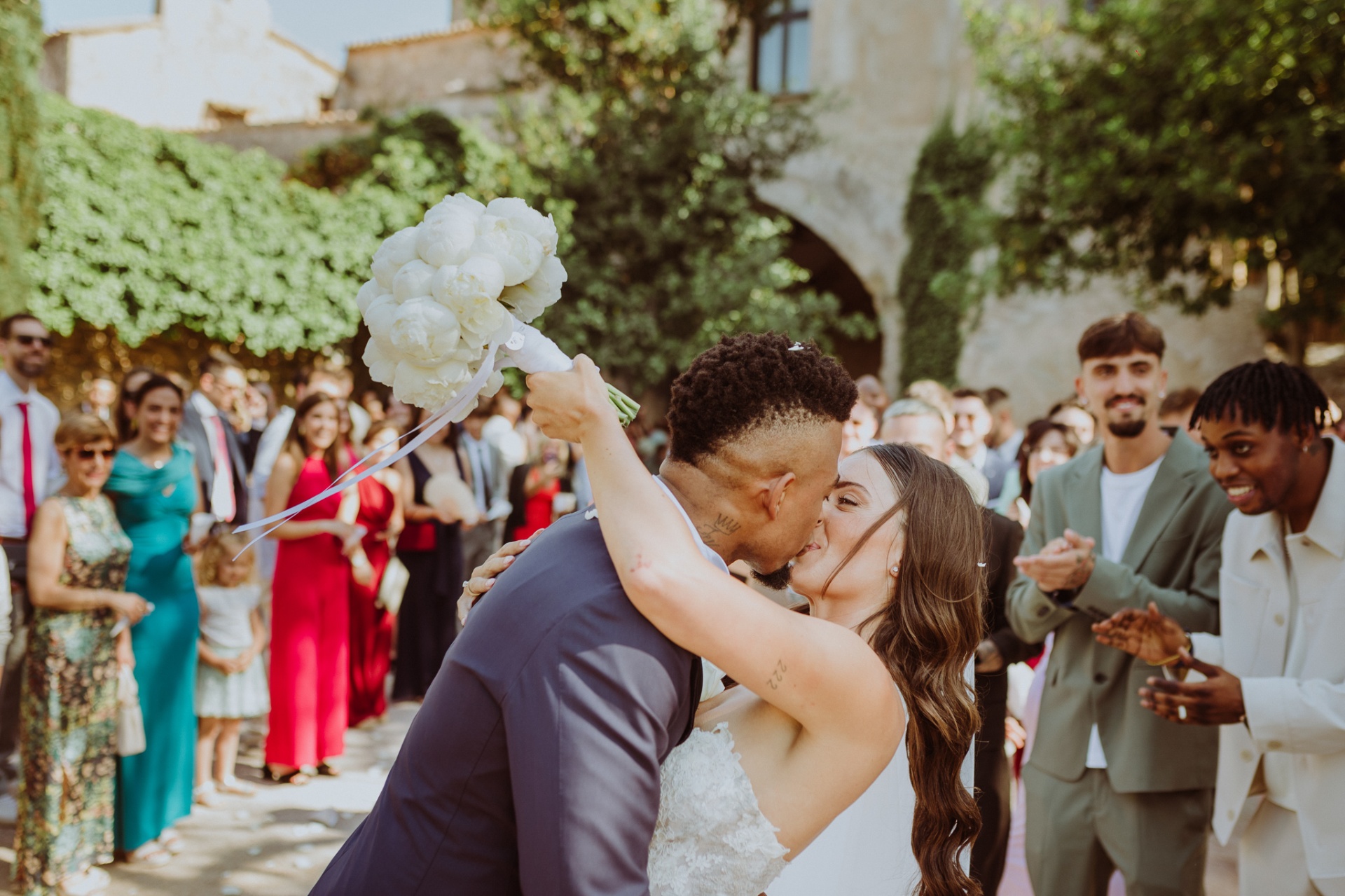 Jose kissing Laura holding peony bouquet high with guests applauding at Castell de Tamarit