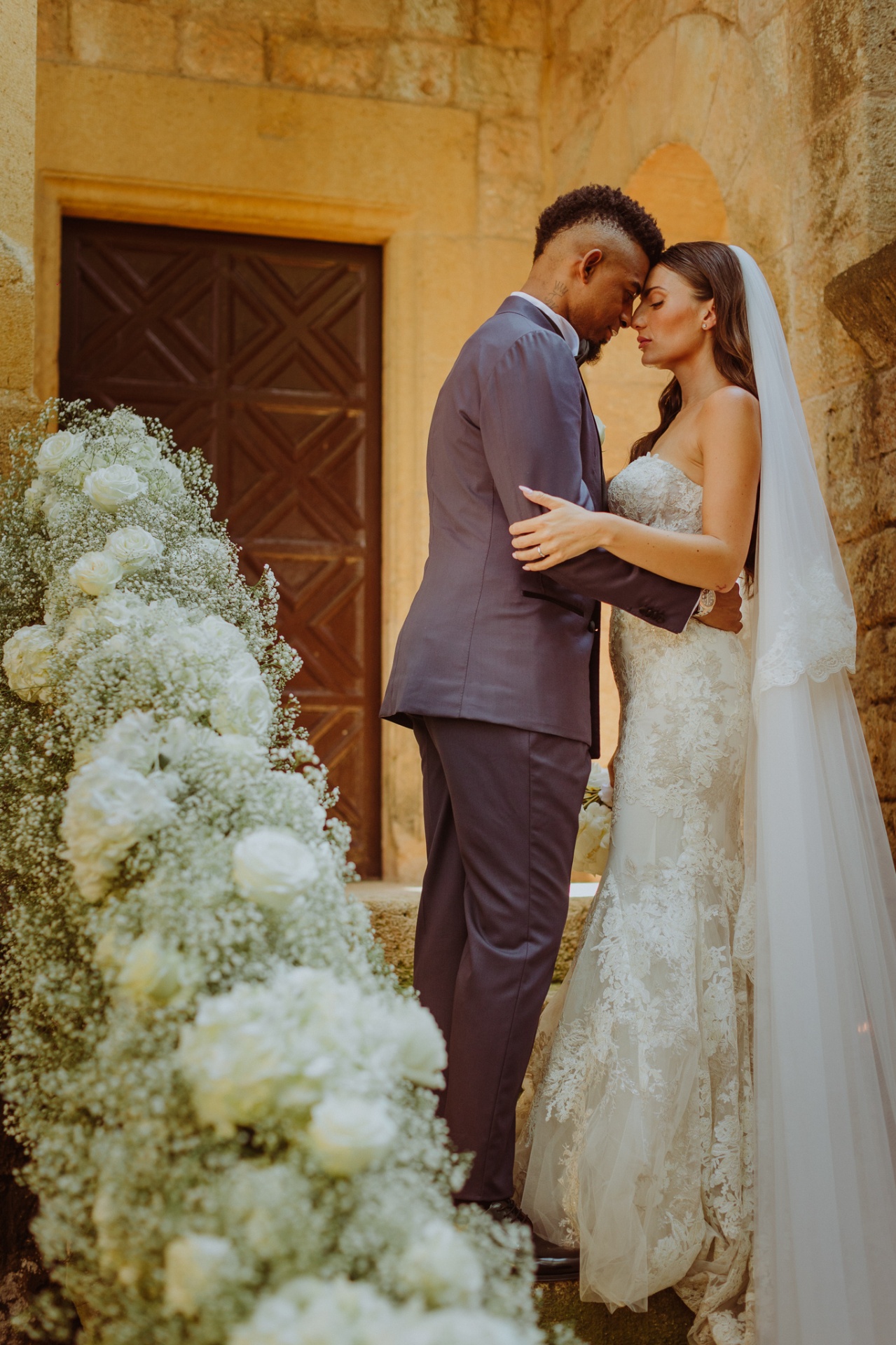 Laura and Jose forehead to forehead surrounded by white roses and baby's breath at castle door