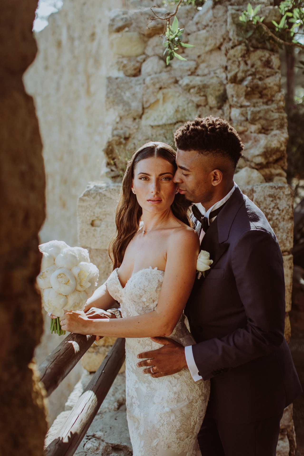 Jose embracing Laura from behind with peony bouquet among castle ruins at golden hour
