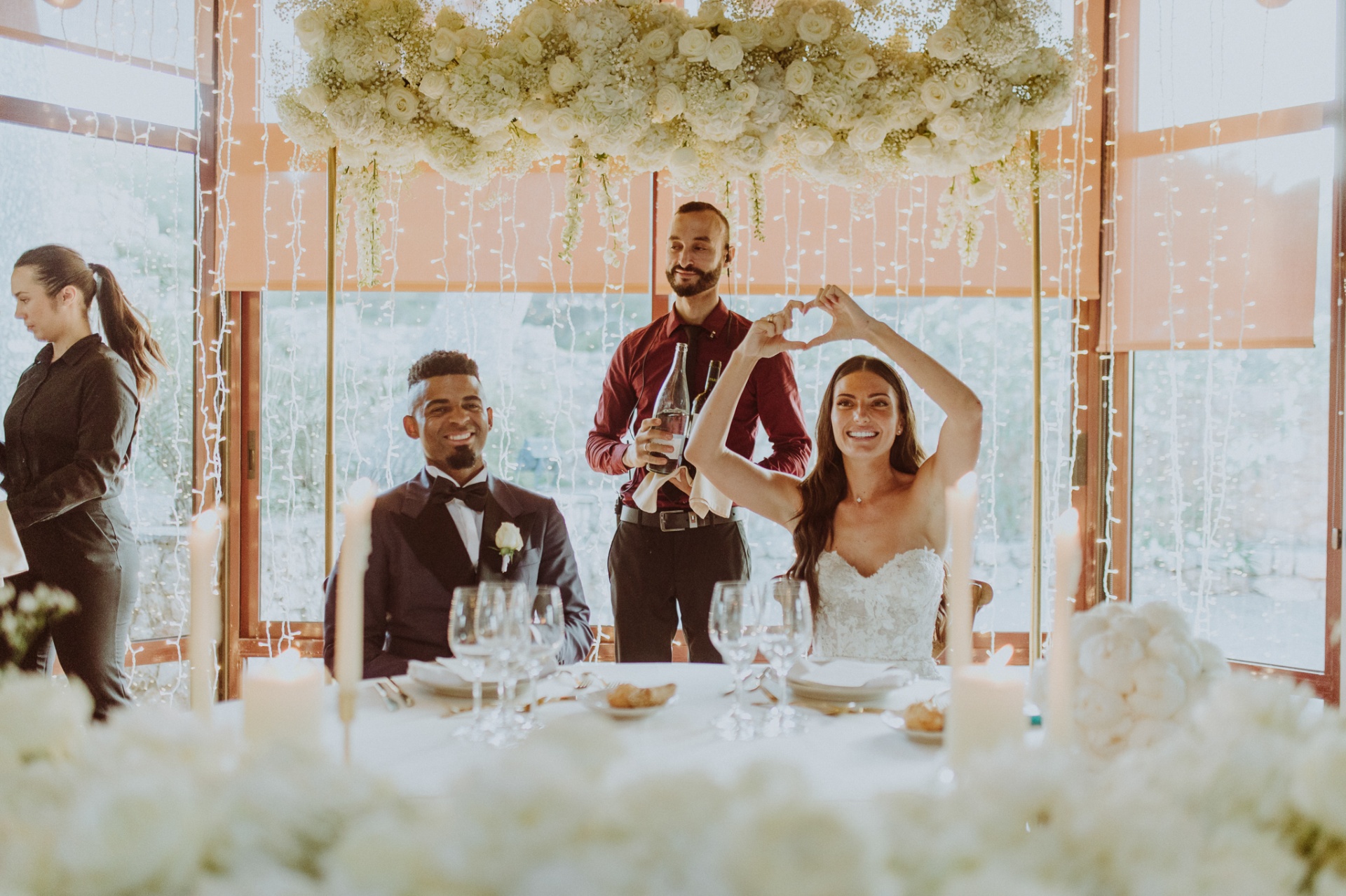 Laura making heart sign with hands at reception table under white floral chandelier with fairy lights