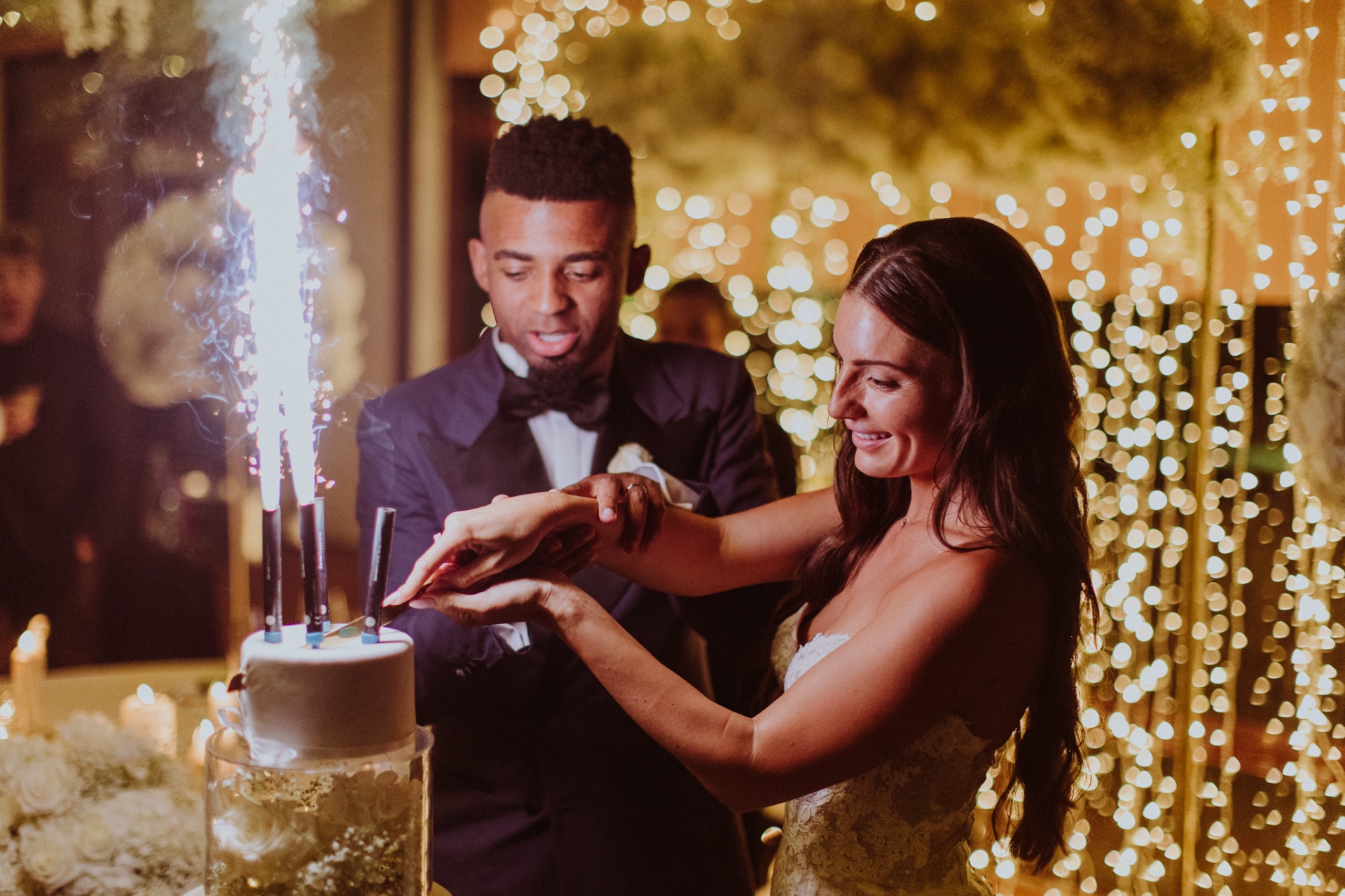 Laura and Jose cutting wedding cake with sparkler fountains and fairy light backdrop