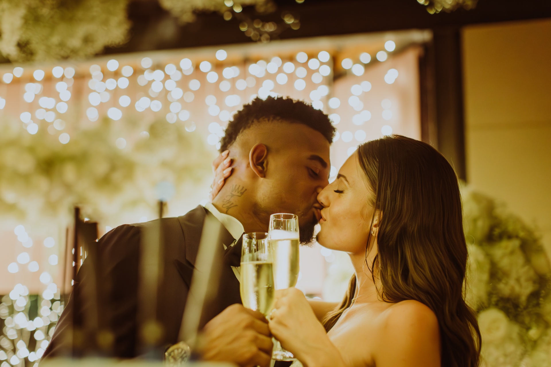 Laura and Jose kissing with champagne glasses under warm bokeh lights at reception