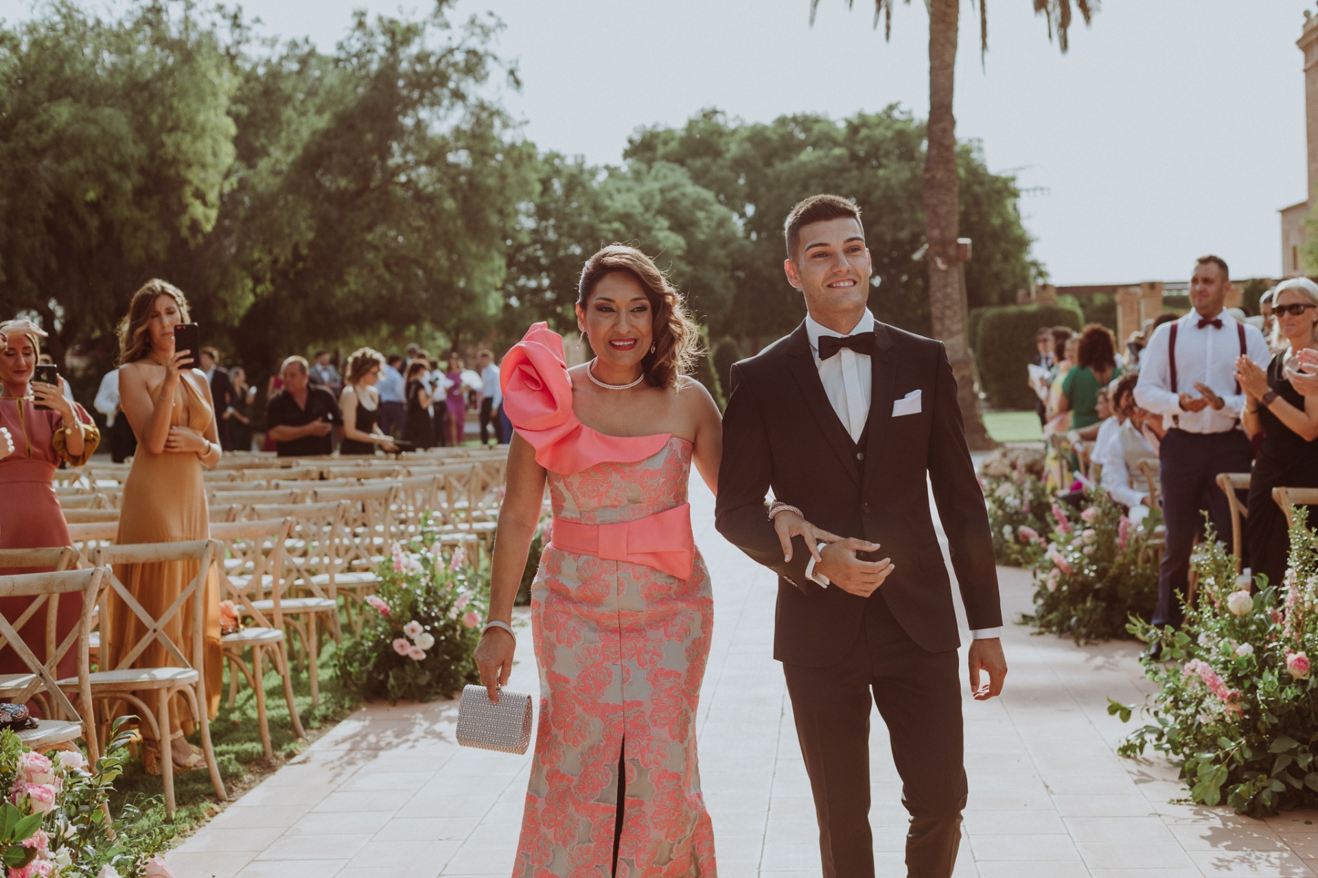 Jorge entrando del brazo de su madre por el pasillo de la ceremonia entre invitados y arreglos florales rosas