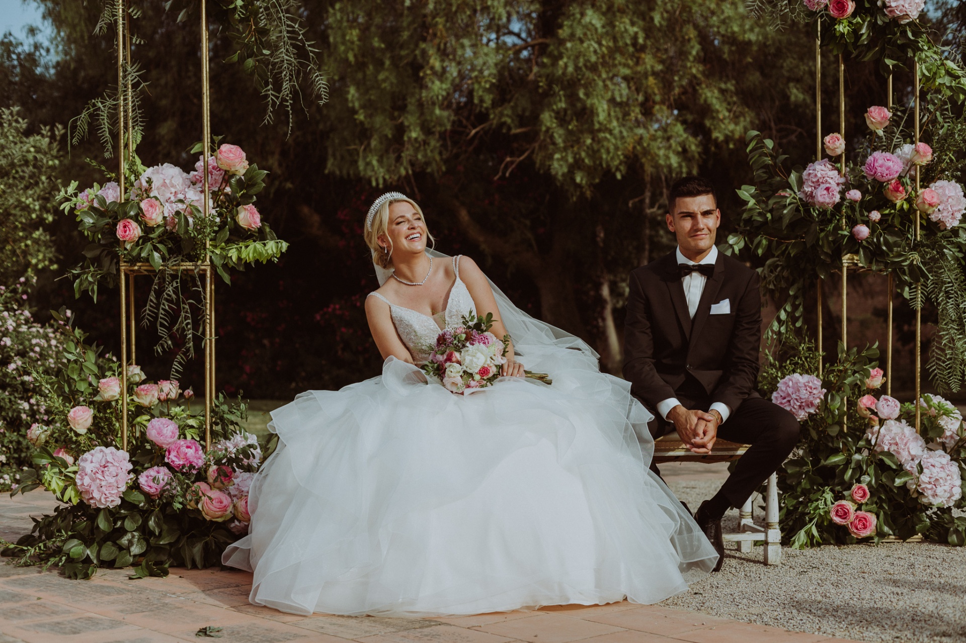 Michelle y Jorge sentados entre arreglos florales rosas y peonías riendo durante la ceremonia en La Cartuja de Ara Christi