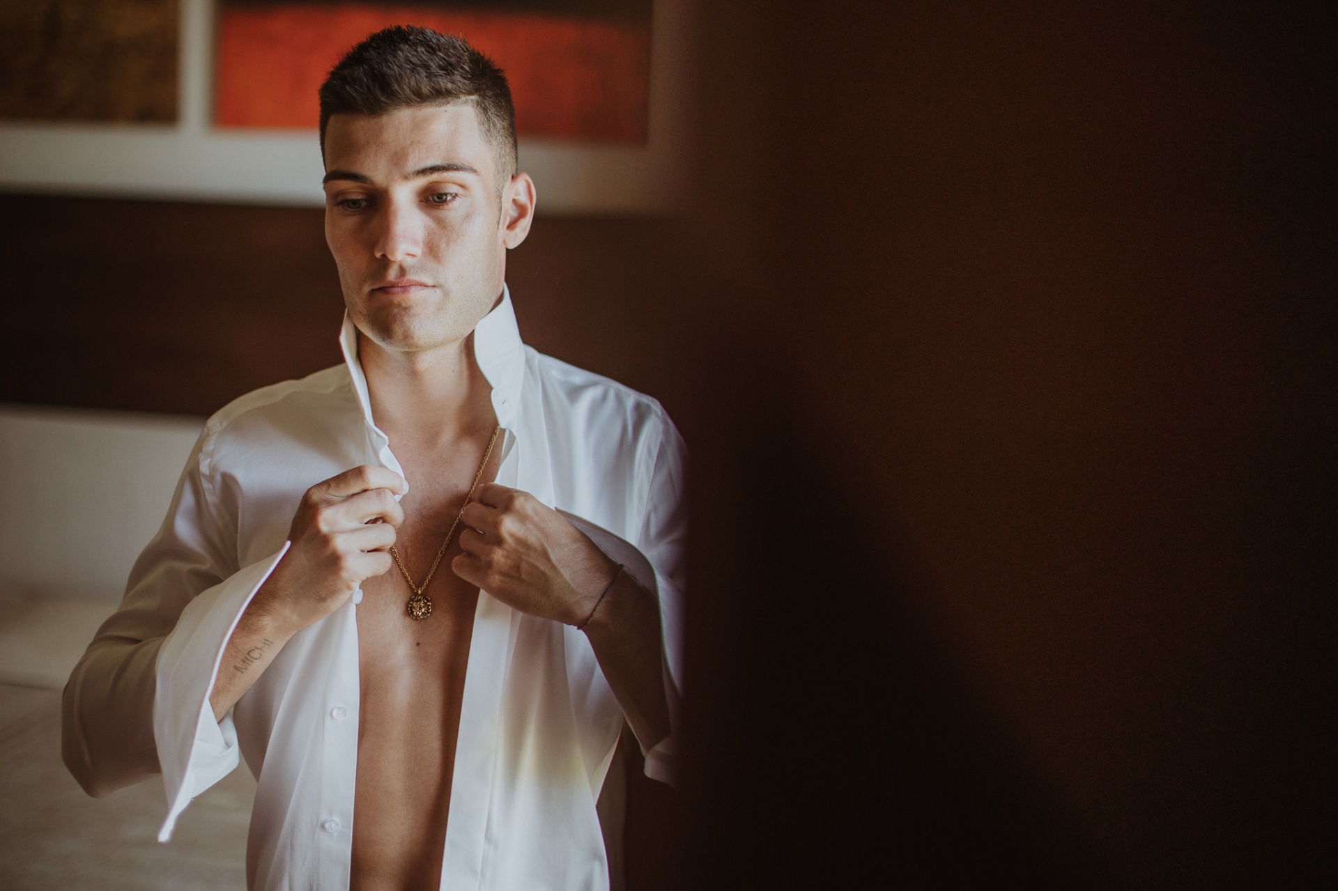 Jorge vistiéndose para la boda, abrochándose la camisa blanca con luz dramática lateral