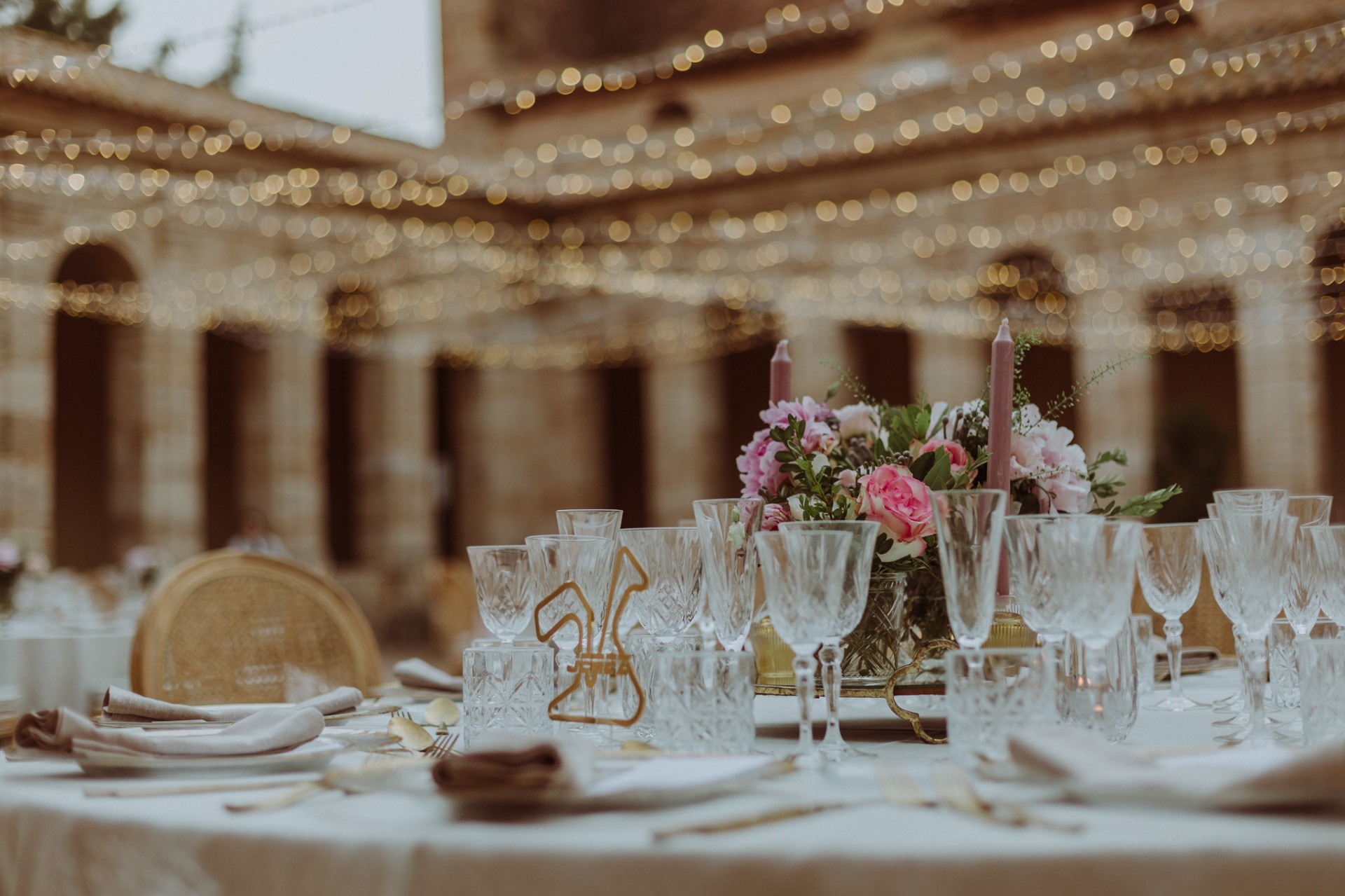 Detalle de la mesa del banquete con cristalería tallada, centro floral de peonías rosas y velas en La Cartuja de Ara Christi