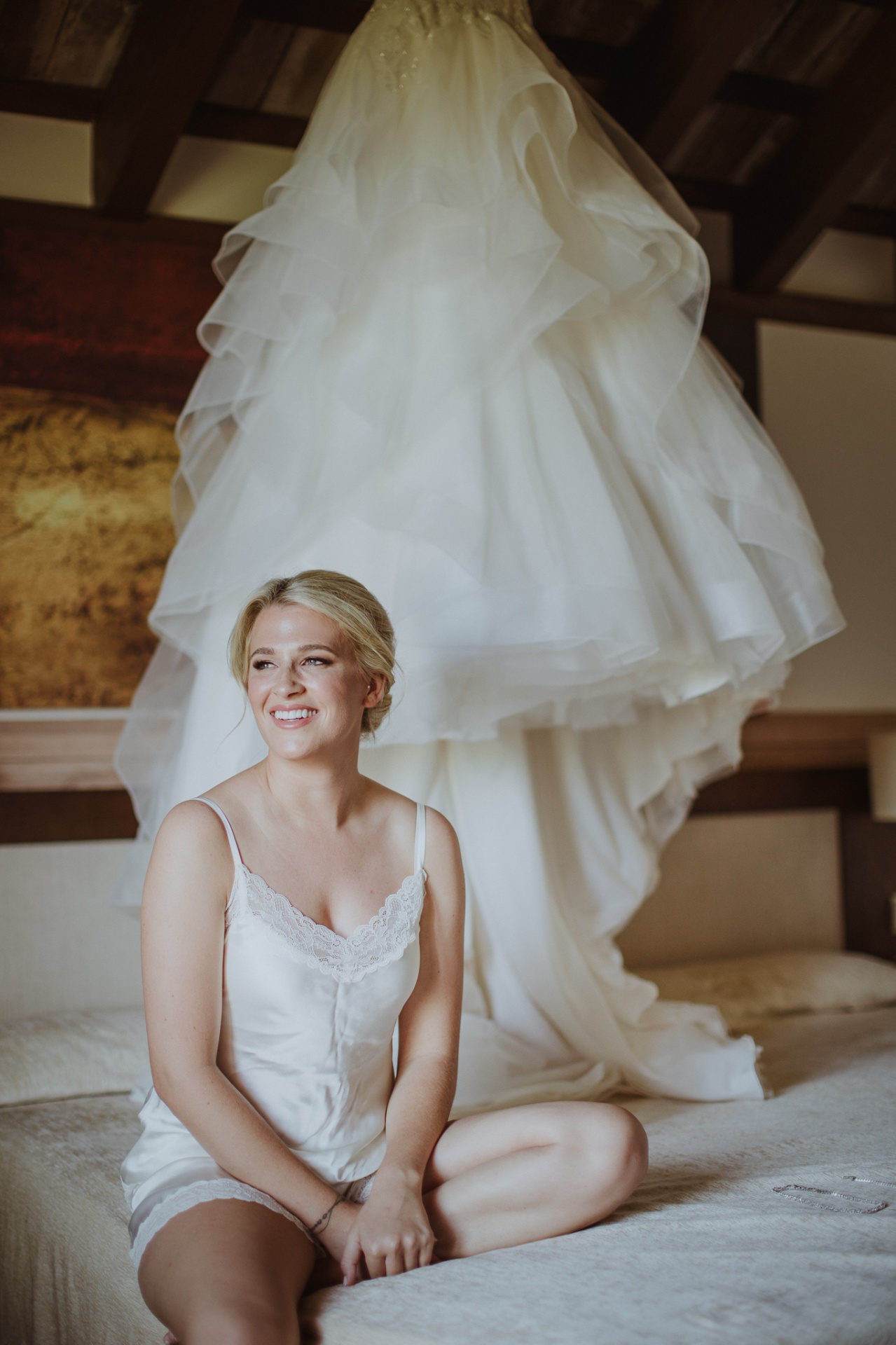 Michelle sonriendo sentada en la cama con su vestido de novia colgado detrás en La Cartuja de Ara Christi